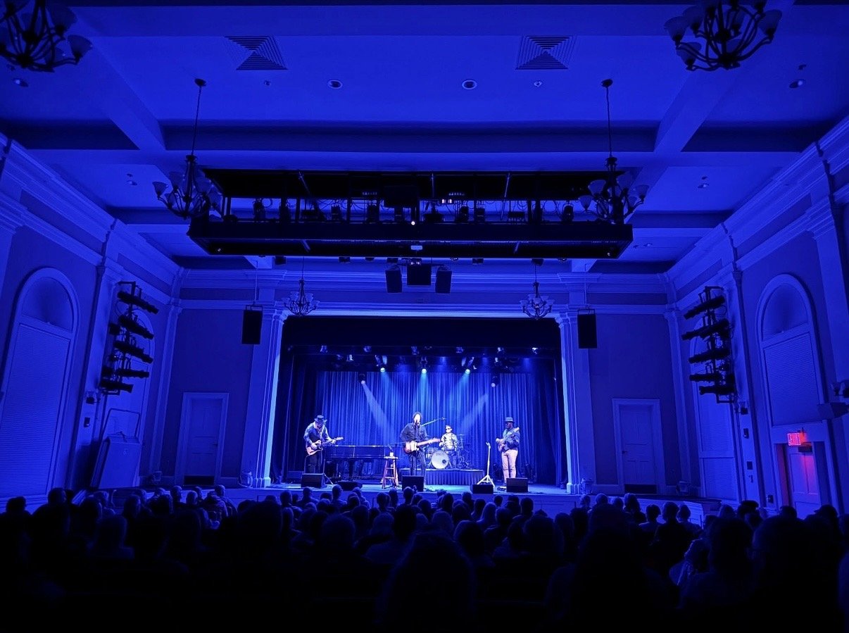 A musical performance on a stage with three performers playing guitars and drums, illuminated by blue stage lights, in an elegant auditorium with an audience watching.