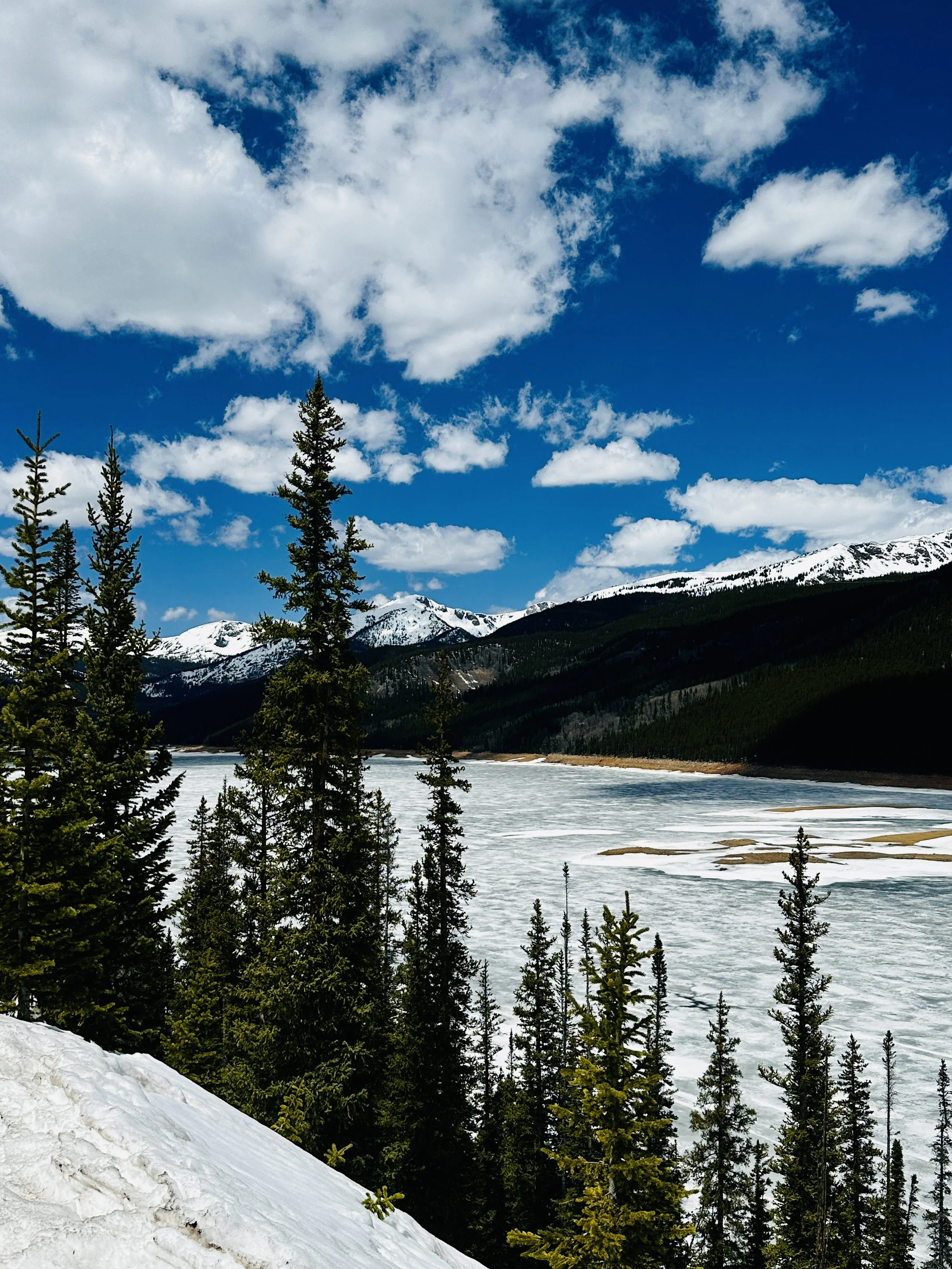 Scenic view of snow-capped mountains, evergreen trees, partly frozen lake, and blue sky with clouds.