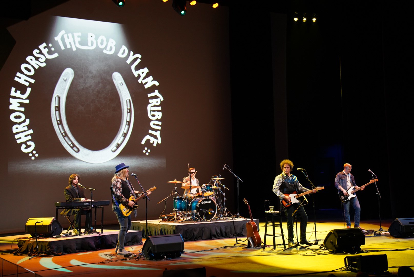 A band performing on stage with a large screen behind them that displays the Horseshoe: The Bob Dylan Tribute logo. The band has five members playing various instruments including keyboard, electric guitar, drums, and bass, with microphones set up fo