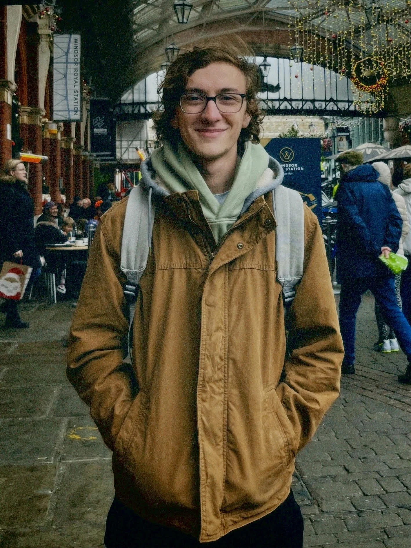 A smiling young man wearing glasses, a brown jacket, a green hoodie, and a gray backpack standing in a covered outdoor market decorated with Christmas lights and wreaths. There are people in the background, some with umbrellas, and a sign indicating Windsor Royal Station.