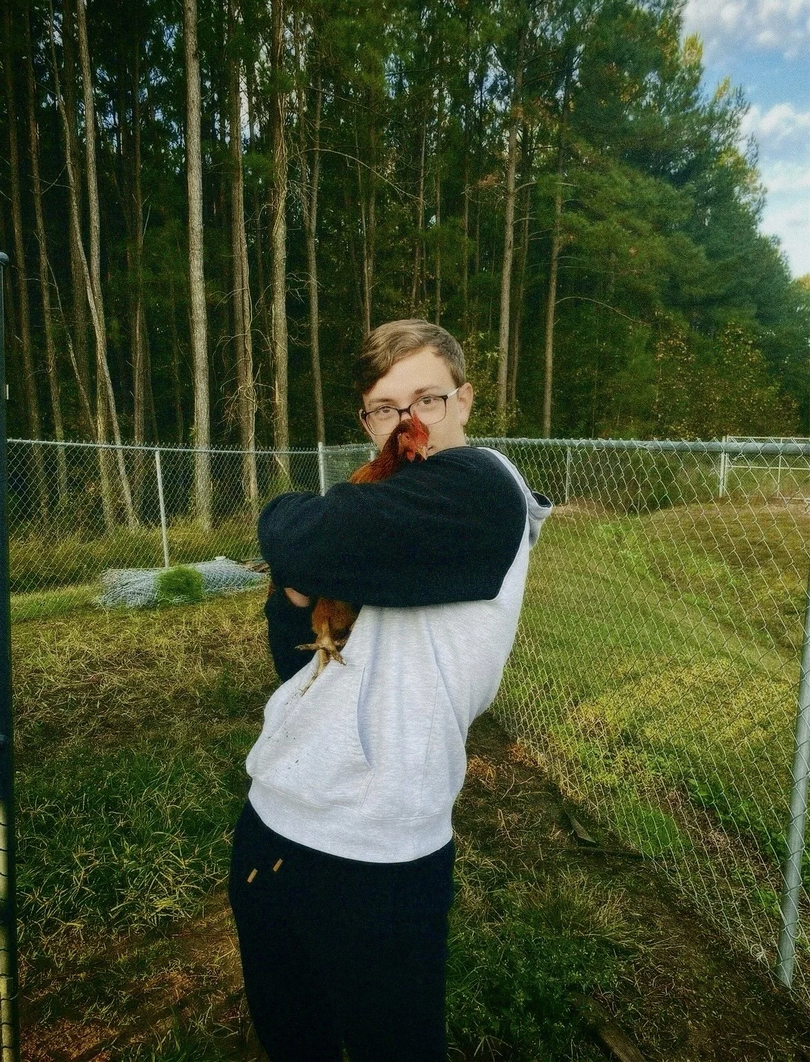A young person with glasses wearing a white hooded sweatshirt and black pants, holding a rooster in front of a chain-link fence and a wooded area with tall trees.