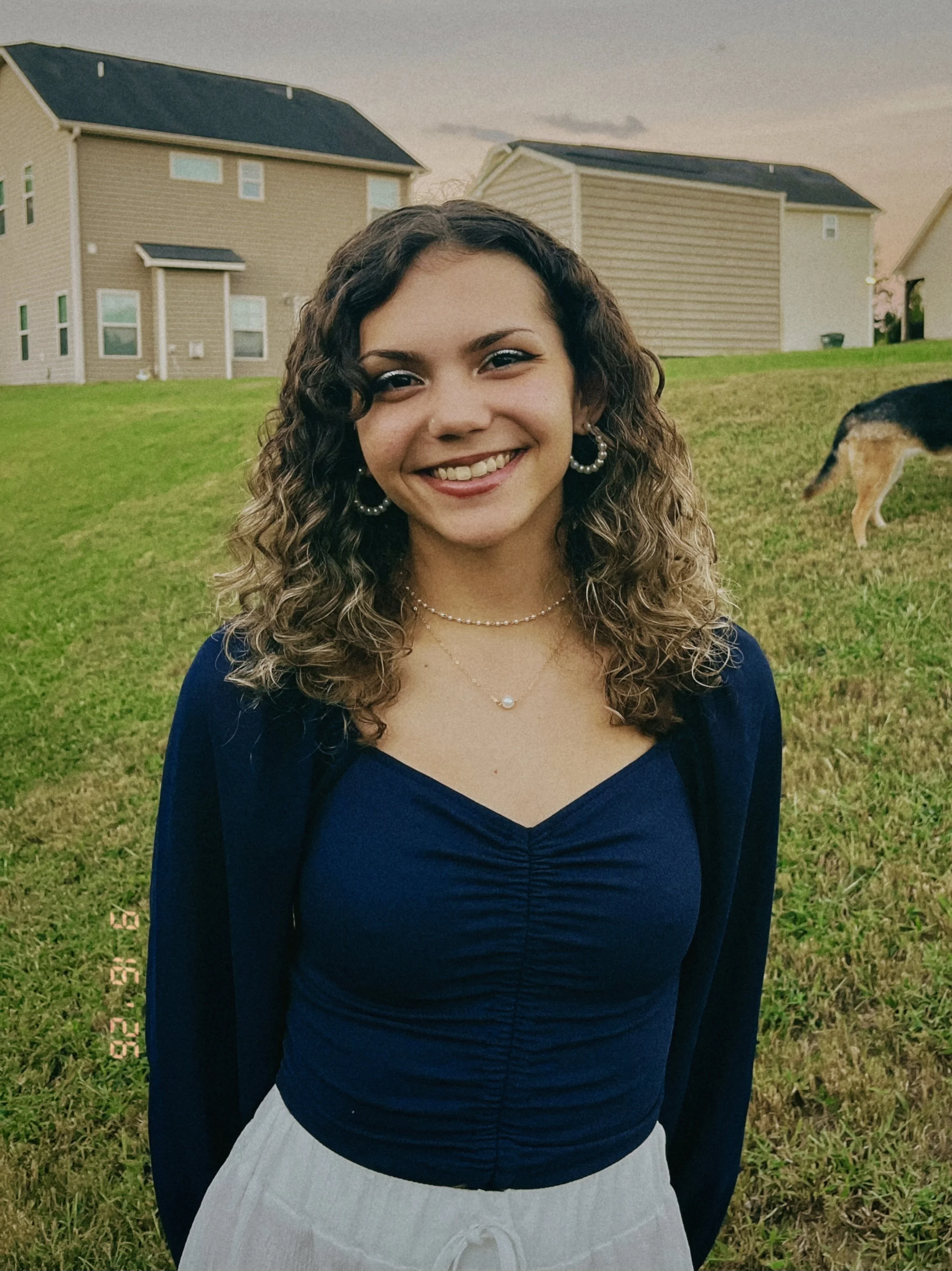 A young woman with curly brown hair, wearing earrings and layered necklaces, smiling outdoors on a grassy area with houses in the background and a dog in the distance.