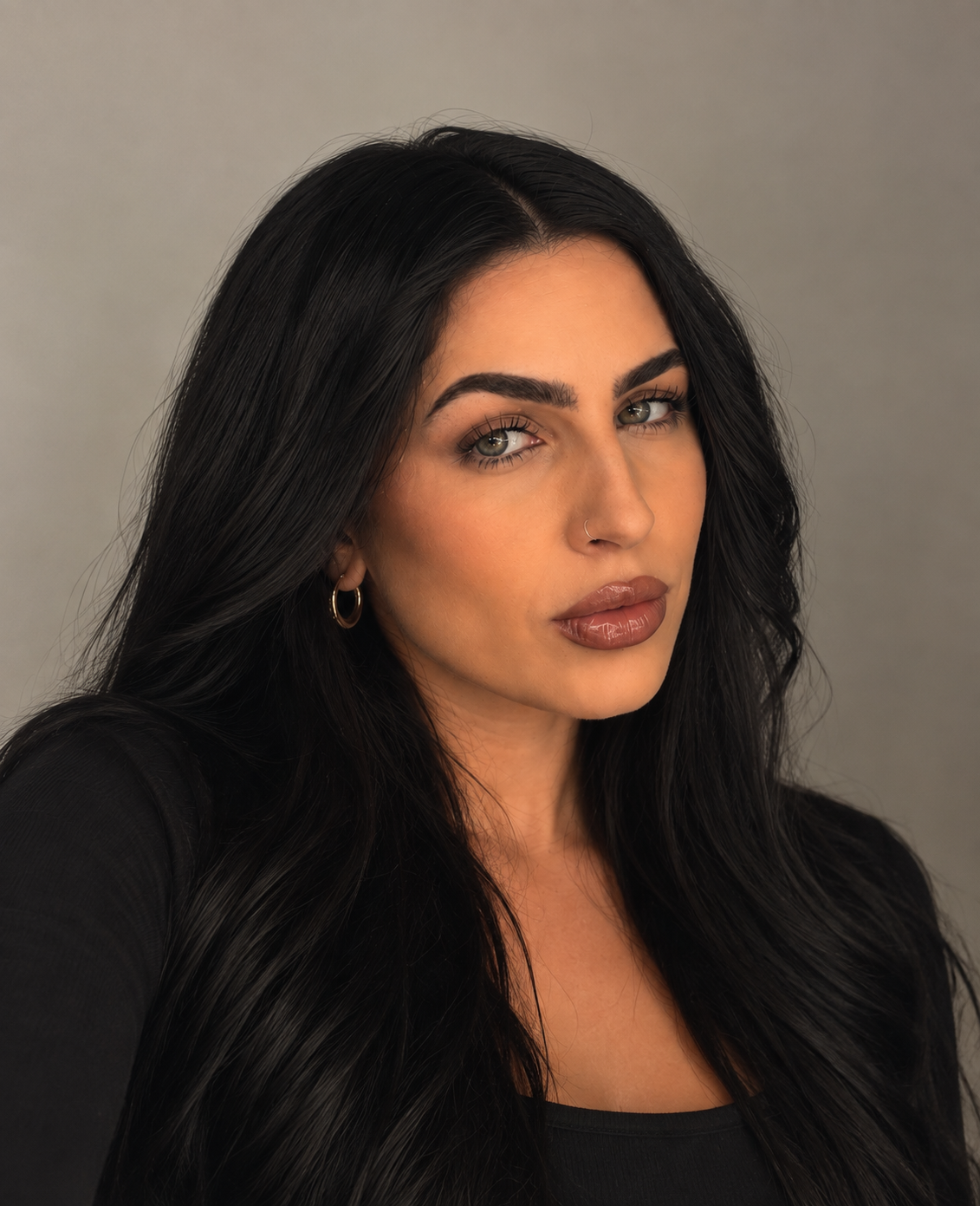 A woman with long black hair, hoop earrings, and makeup, wearing a black top, posing against a plain gray background.
