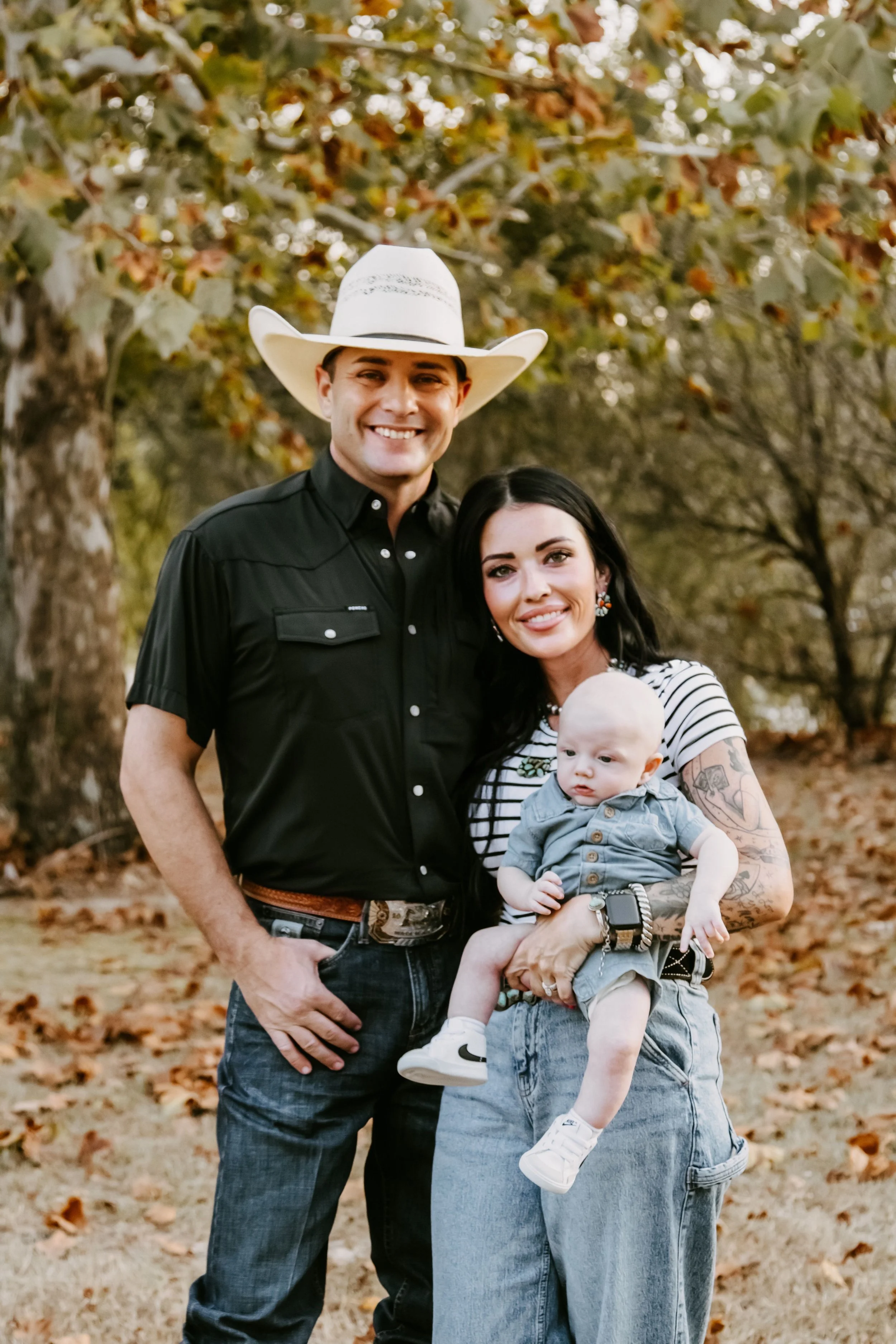 A smiling man wearing a white cowboy hat and a black western shirt standing outdoors, with a woman with tattoos and a black and white striped shirt holding a baby in front of trees with autumn leaves.