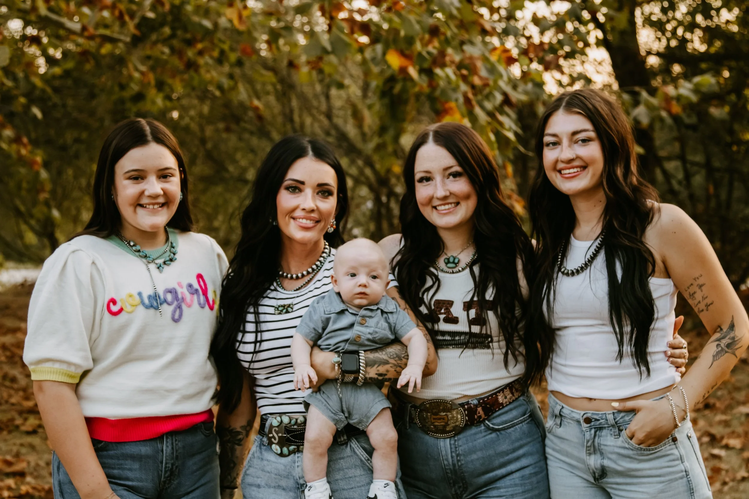 A group of five women and one baby, smiling outdoors in a park with autumn trees in the background.