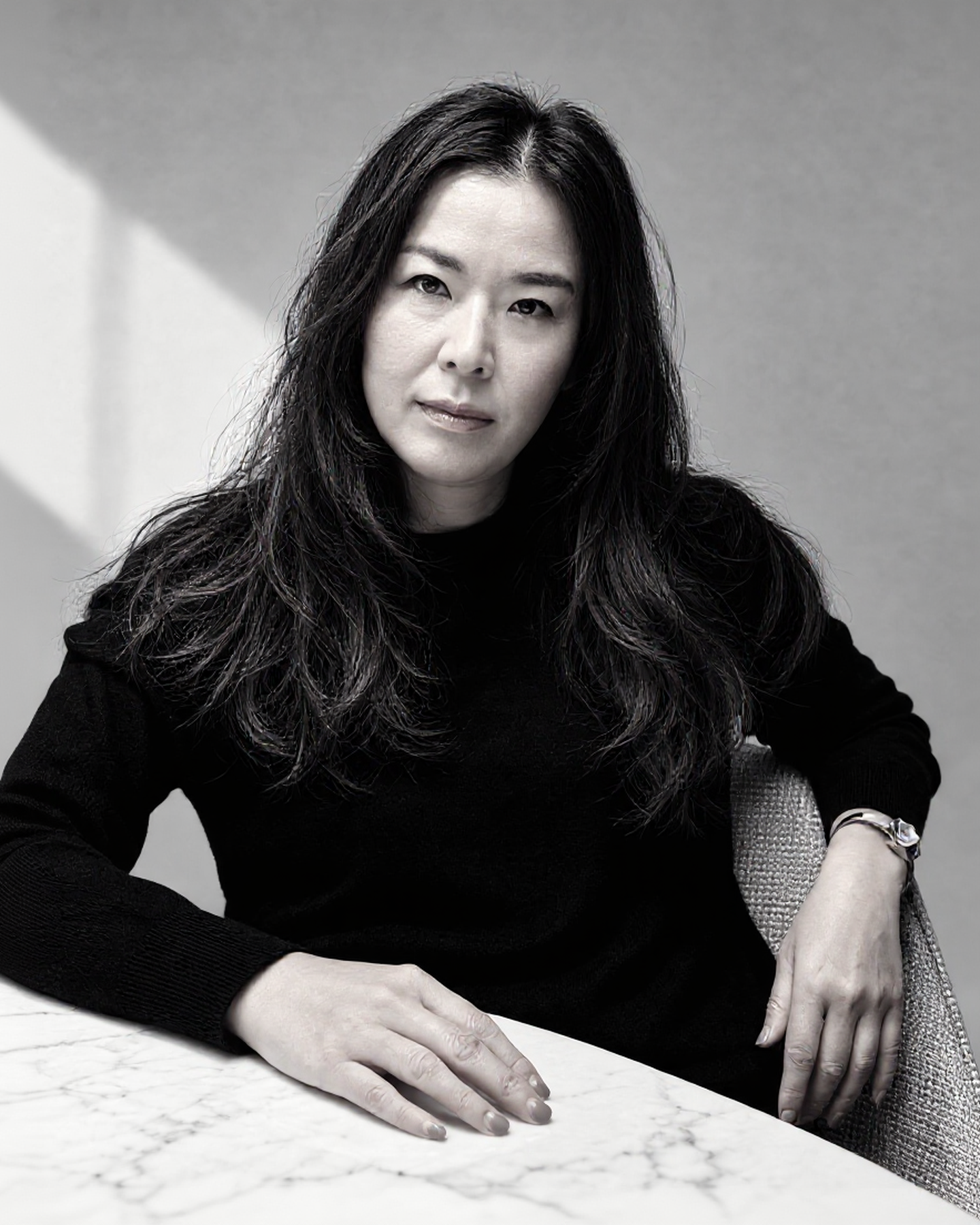 Black and white portrait of a woman with long, dark, wavy hair, sitting at a marble table, wearing a black top, with a grey background.