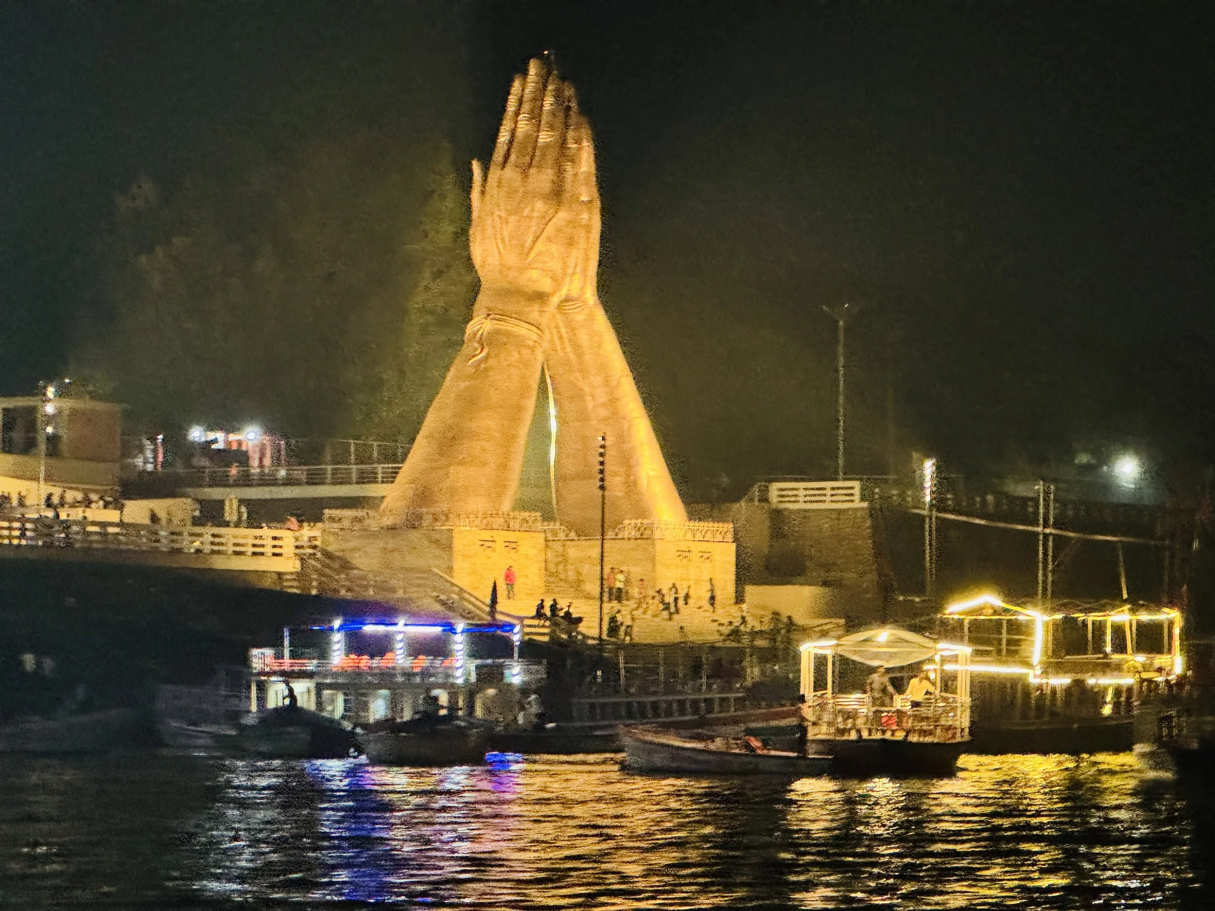 Nighttime waterfront view of a large illuminated praying-hands monument surrounded by boats and colorful reflections on the water
