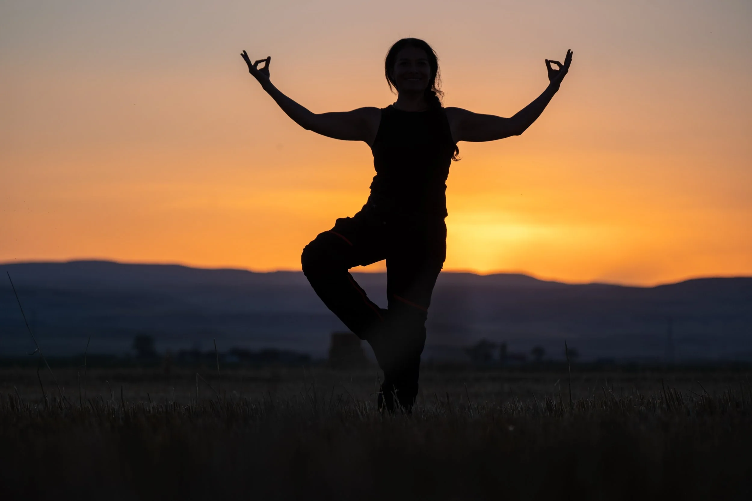 Silhouette of a person practicing yoga or meditation in the tree pose during sunset, with colorful sky in the background.