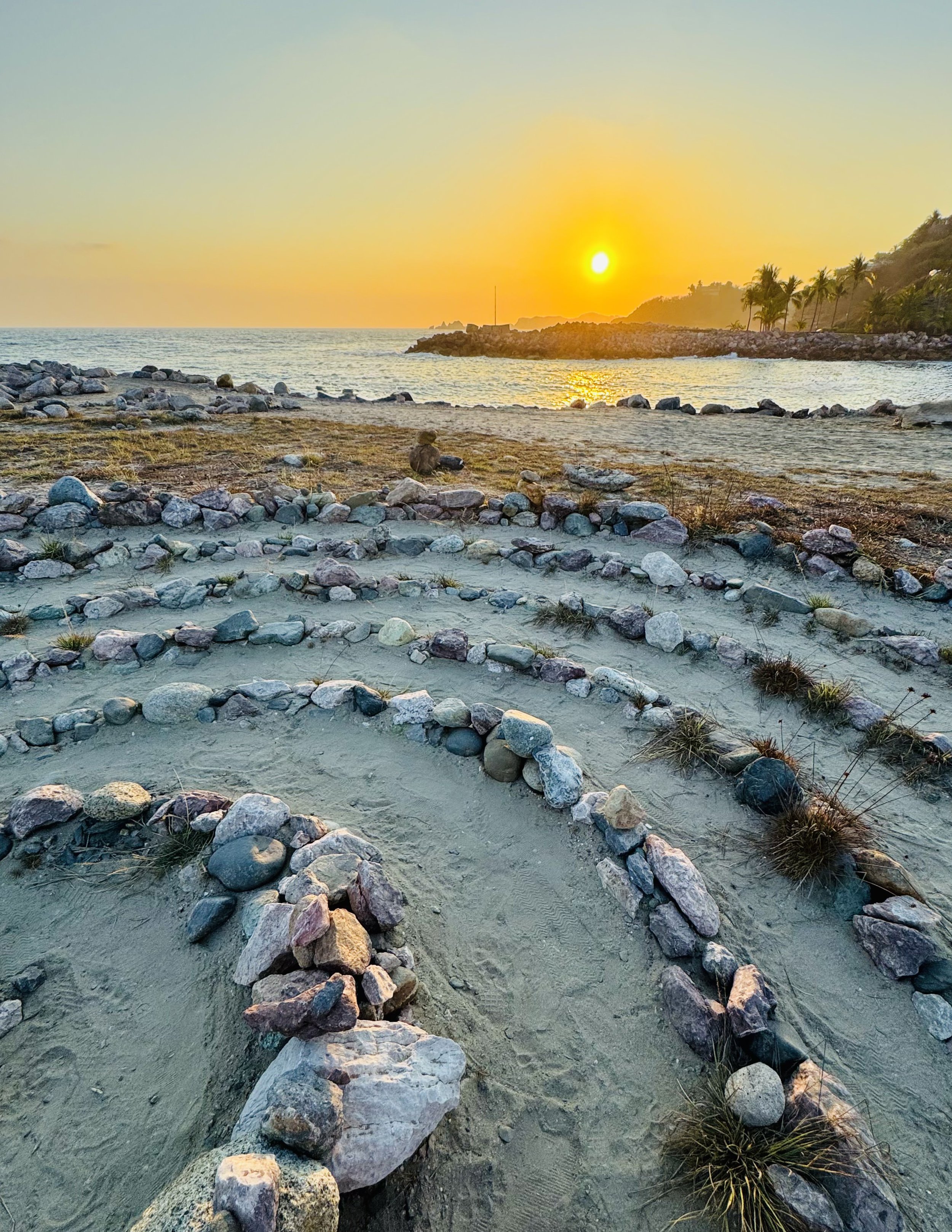 Rocky tropical shoreline at sunset with calm ocean water, palm trees, and a sunlit hillside.