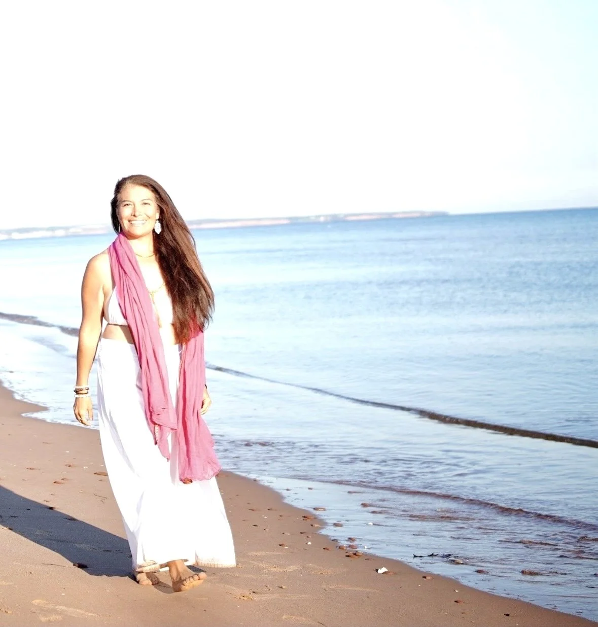 Smiling woman in a white dress and pink scarf standing on a sunny beach beside the ocean