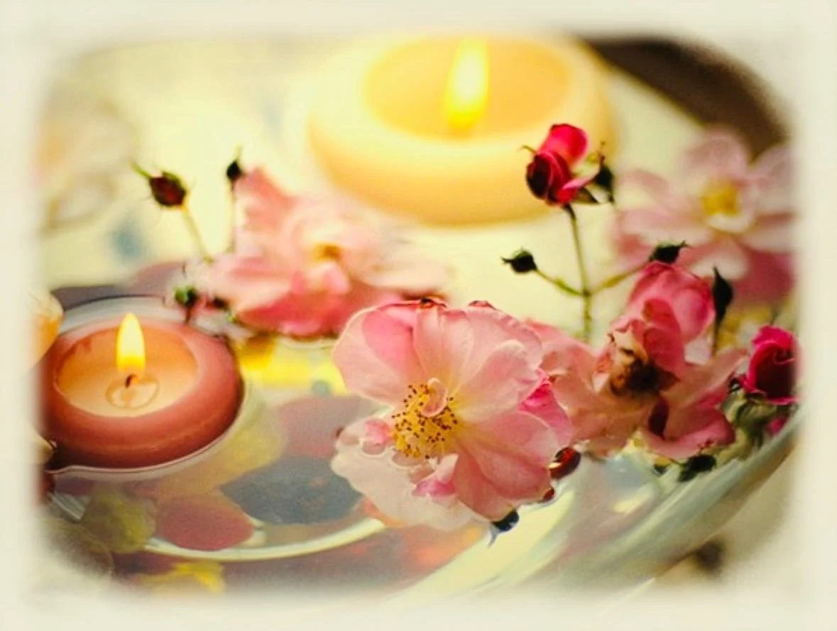 Pink flowers and rosebuds arranged around two glowing candles in a soft pastel still life