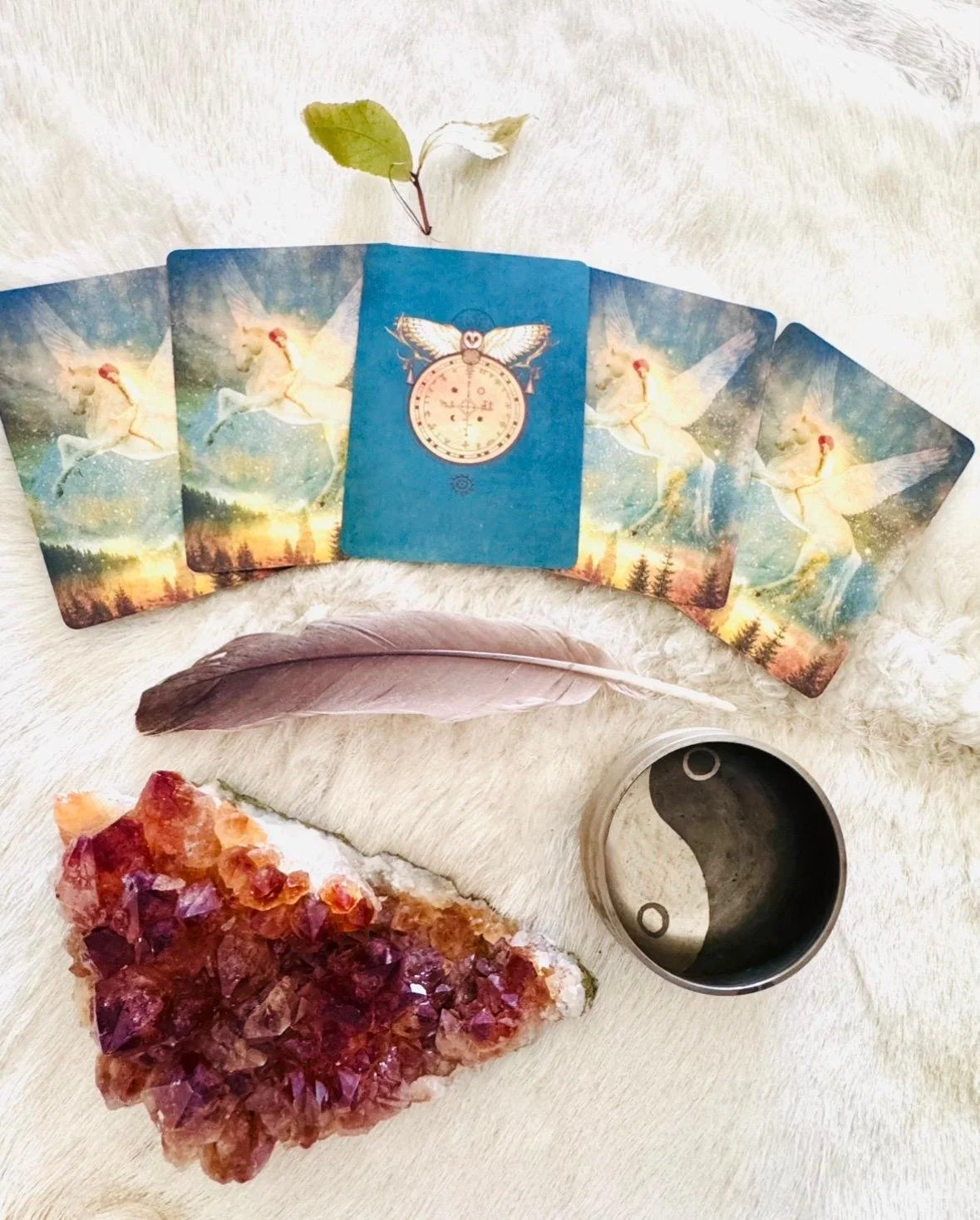 Spiritual altar with tarot cards, feather, leaf, yin-yang dish, and purple crystal cluster on white fabric