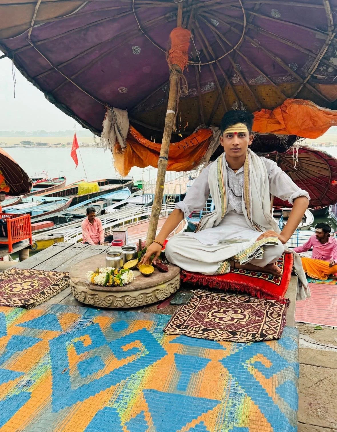 Young Hindu priest seated beneath a decorated canopy on a boat-covered riverbank, with offerings and colorful boats in the background