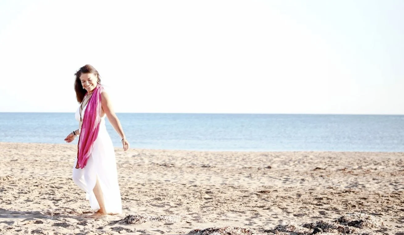 Woman in a white dress and pink scarf walking along a sunny sandy beach beside the ocean