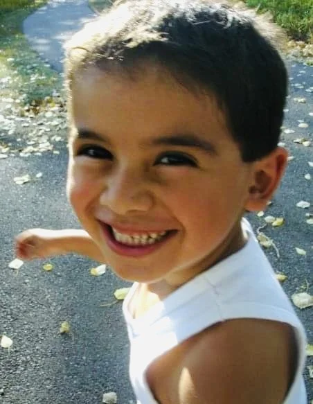 Smiling young boy in a white sleeveless shirt outdoors on a leaf-strewn path