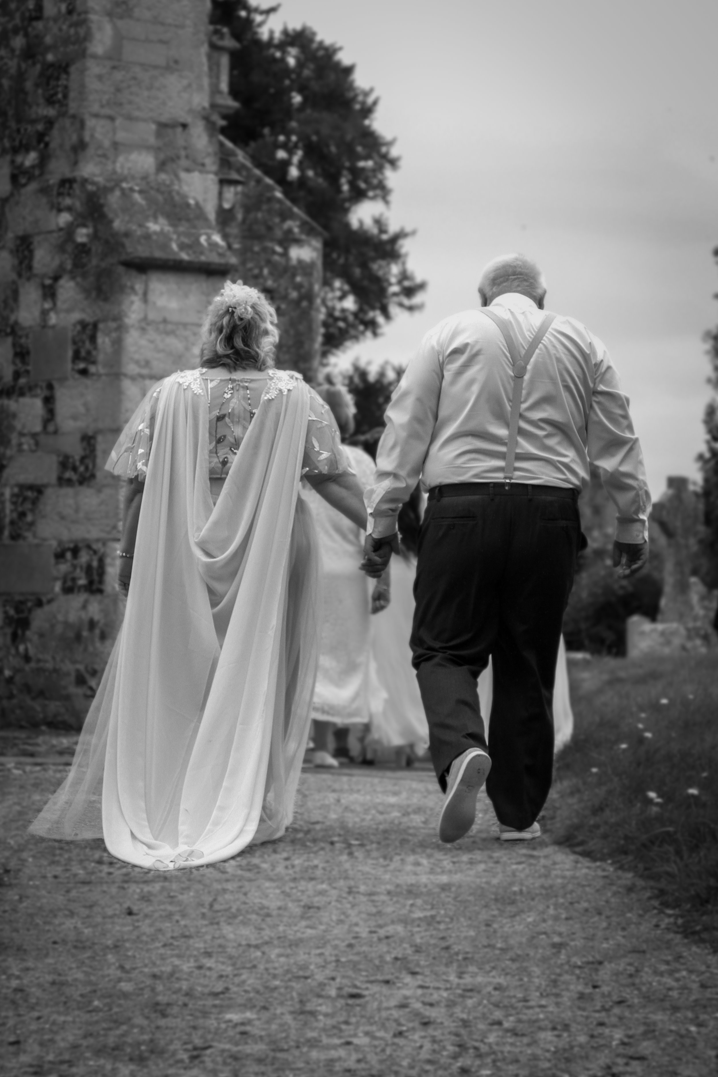 An elderly couple walking hand in hand outdoors at dusk, with a stone building and trees in the background.