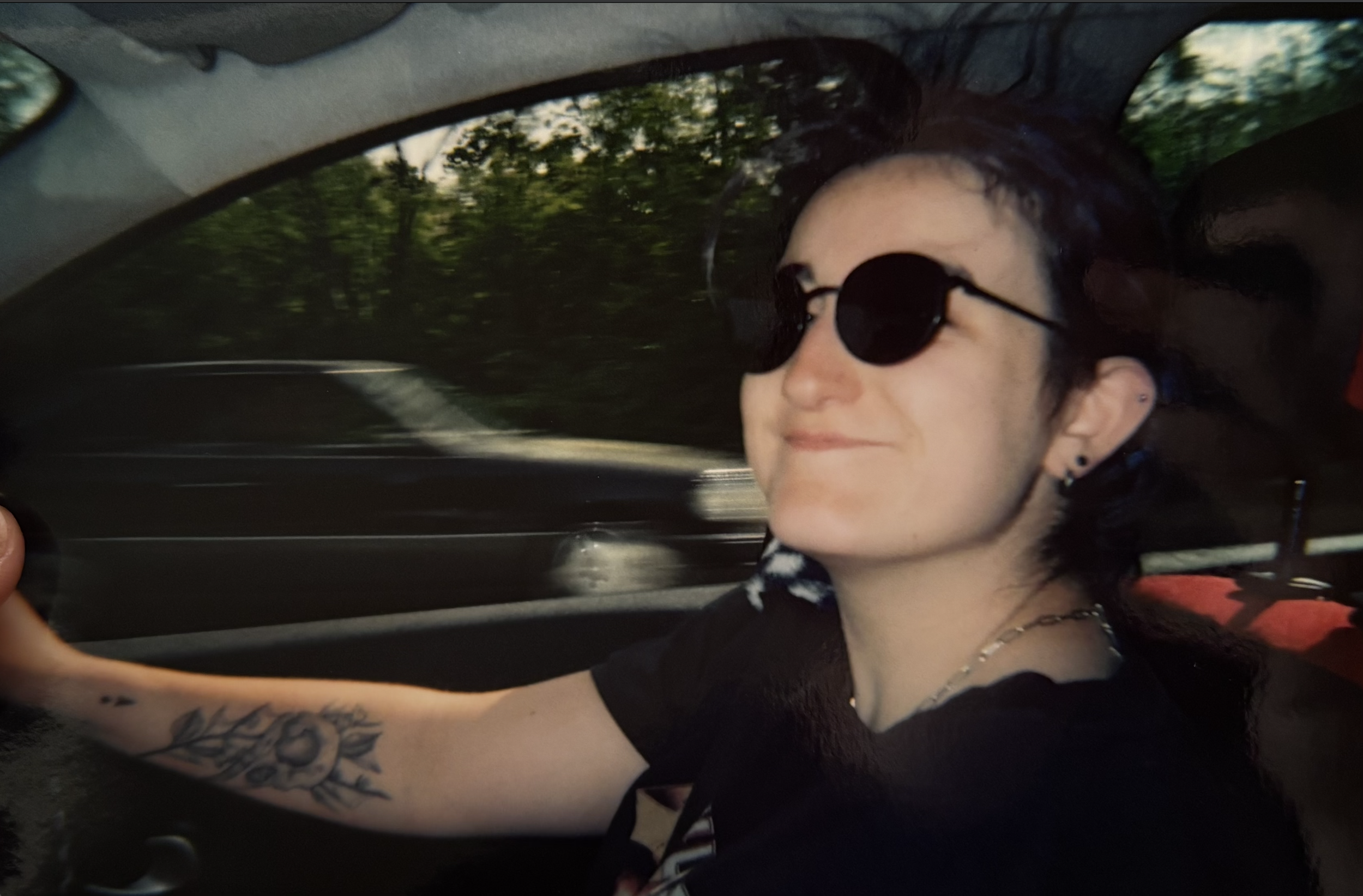 A young woman with dark hair, wearing sunglasses, a black shirt, and earrings, taking a selfie while driving in a car.