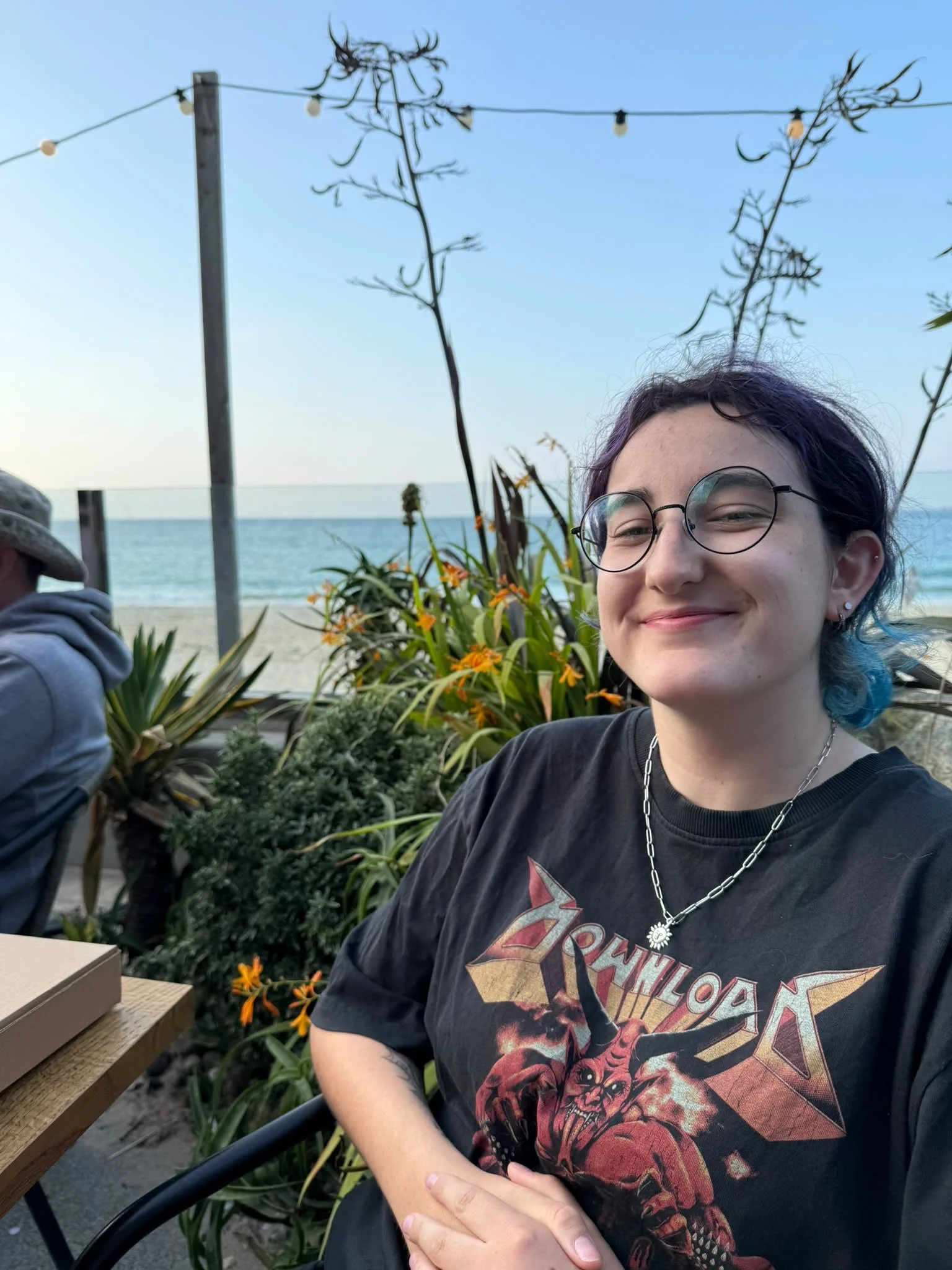 A smiling person with glasses and purple-blue hair at the beach, wearing a black T-shirt with a graphic, surrounded by orange flowers and green plants, with the ocean and sky in the background.