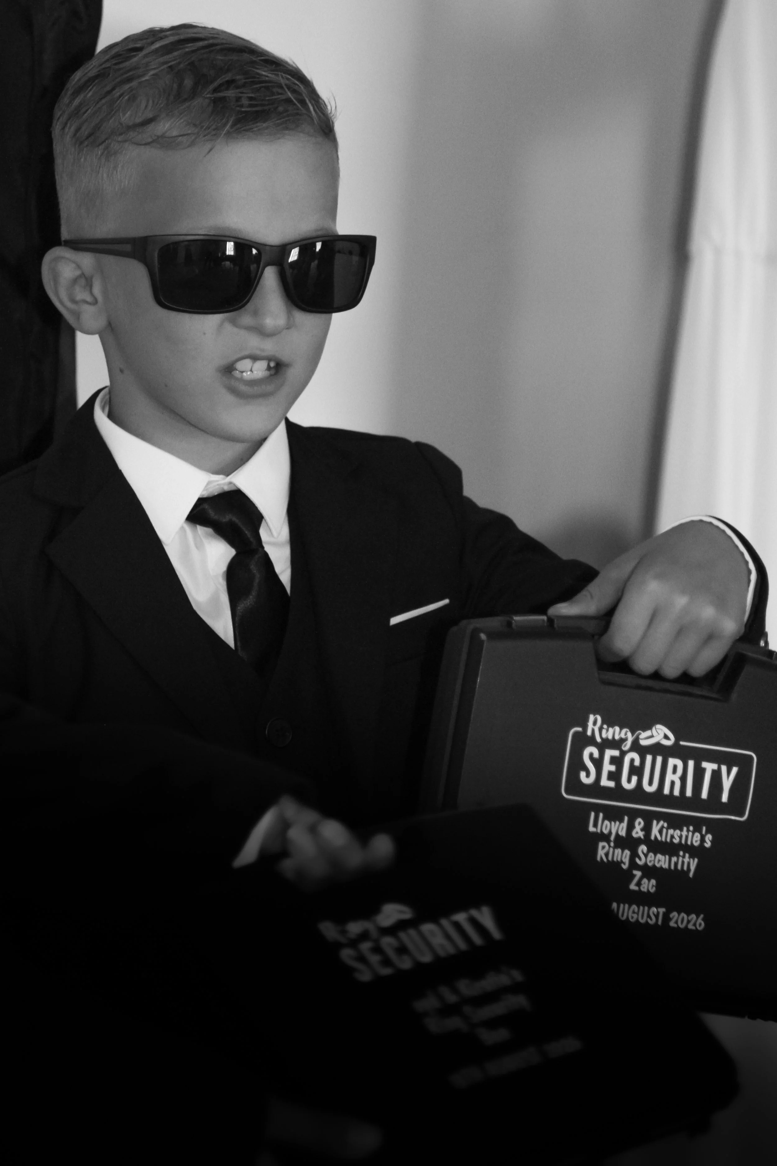 Young boy wearing sunglasses, a suit, and a tie, sitting at a table with a sign that reads 'Ring Security Lloyd & Kirstie's Ring Security Zac August 2026'.