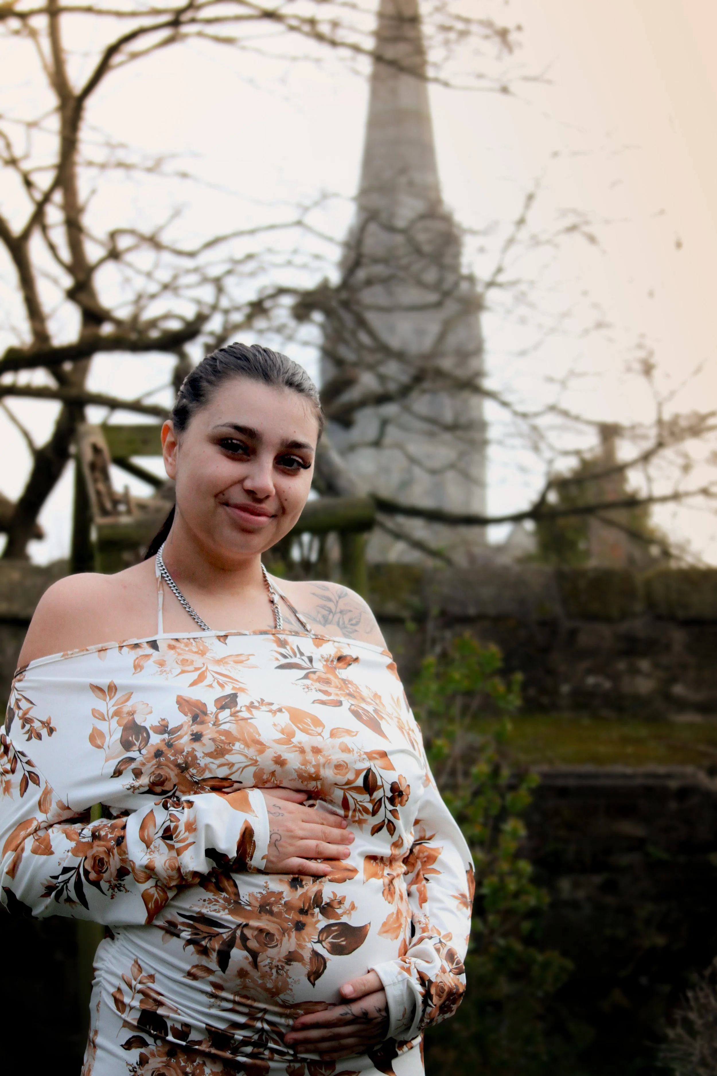 A pregnant woman with dark hair tied back, wearing an off-the-shoulder floral dress, holding her belly, standing outdoors with an Eiffel Tower replica in the background and leafless trees.