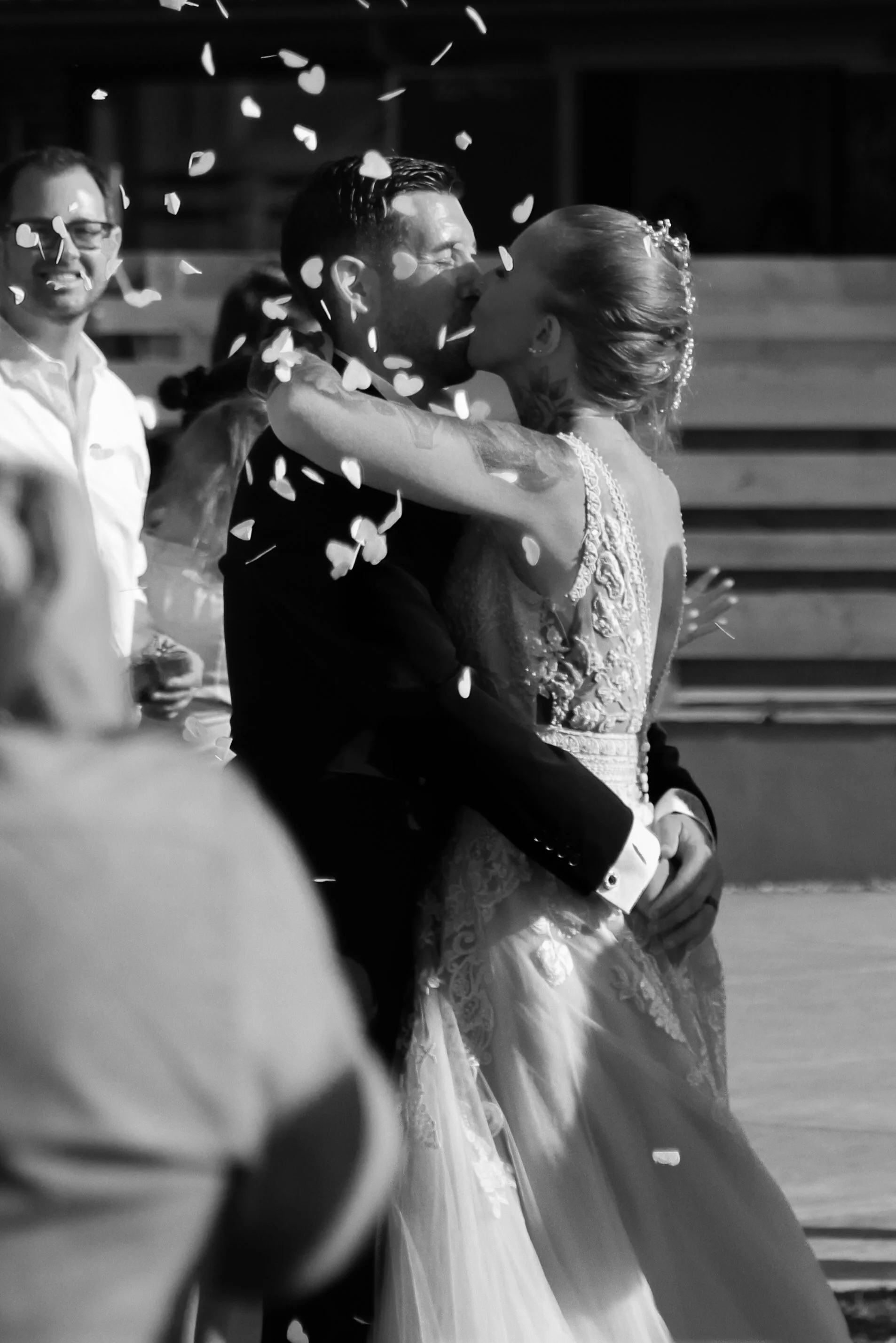 A black and white photo of a bride and groom kissing during a wedding ceremony, with guests and confetti in the background.