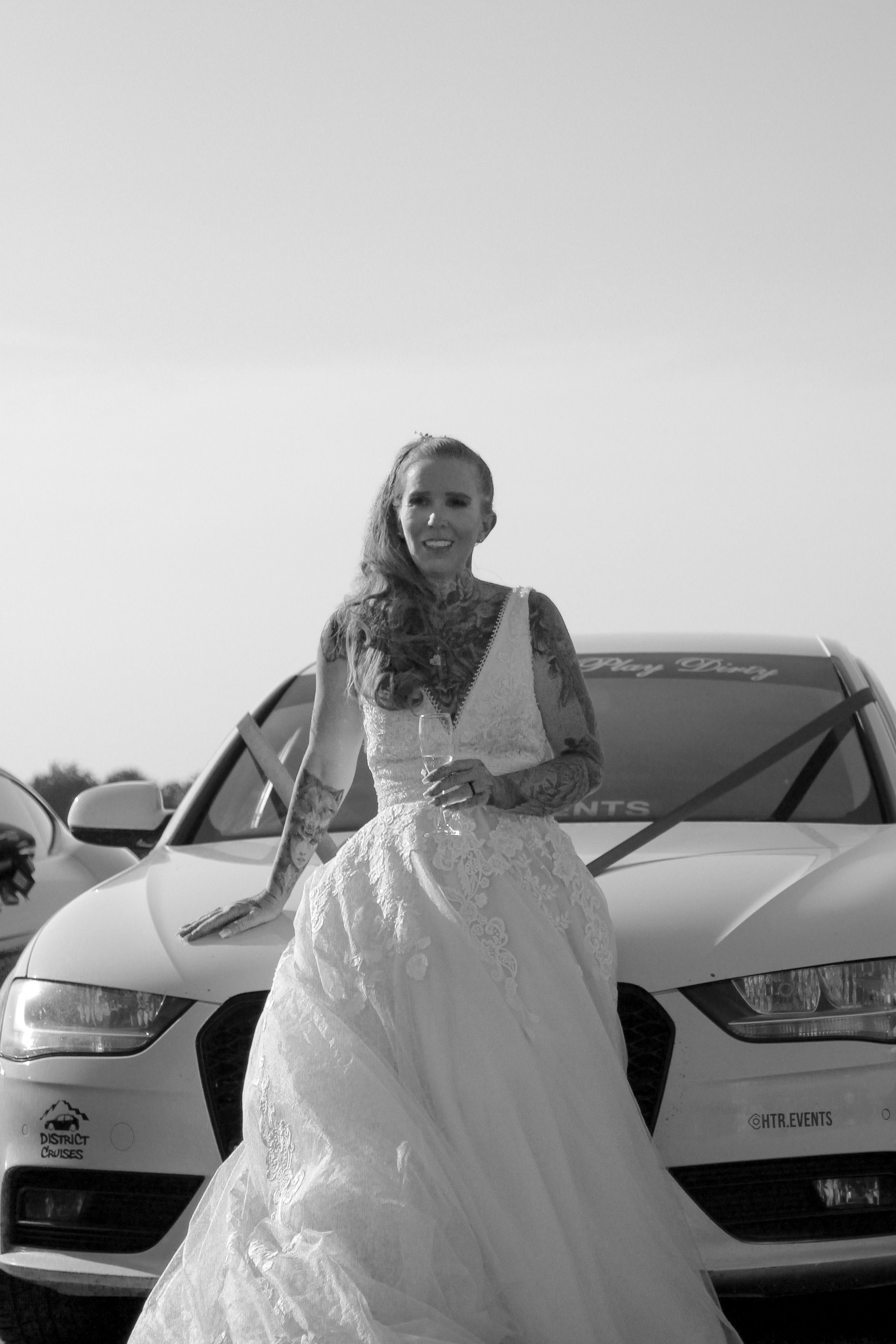 A woman in a white lace wedding dress holding a glass of wine, standing in front of a white car in an outdoor setting.