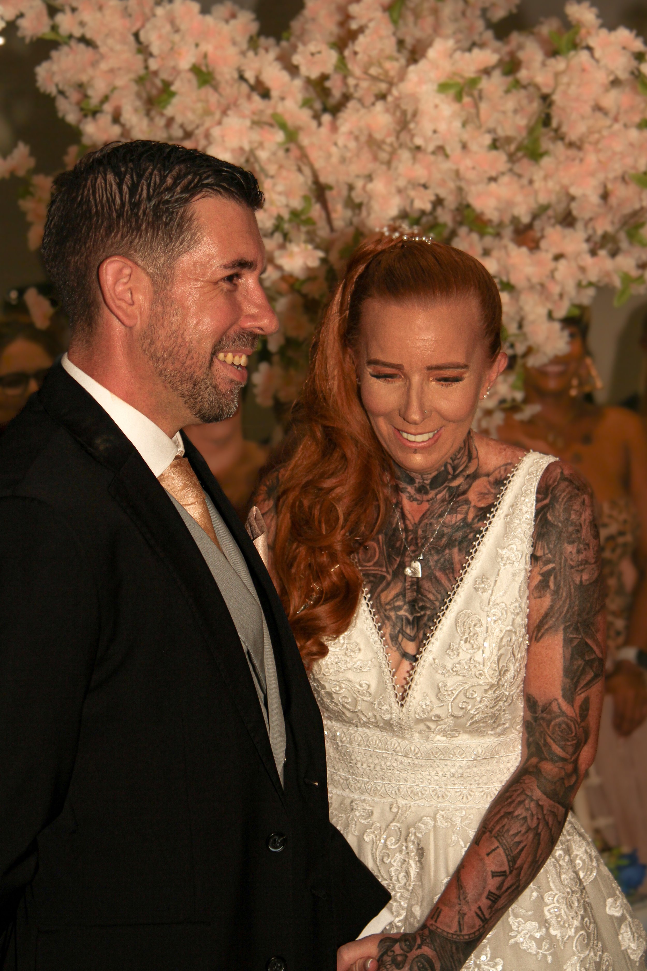 A couple dressed in wedding attire holding hands, with a background of pink flowers and blurred guests, smiling and sharing a special moment.