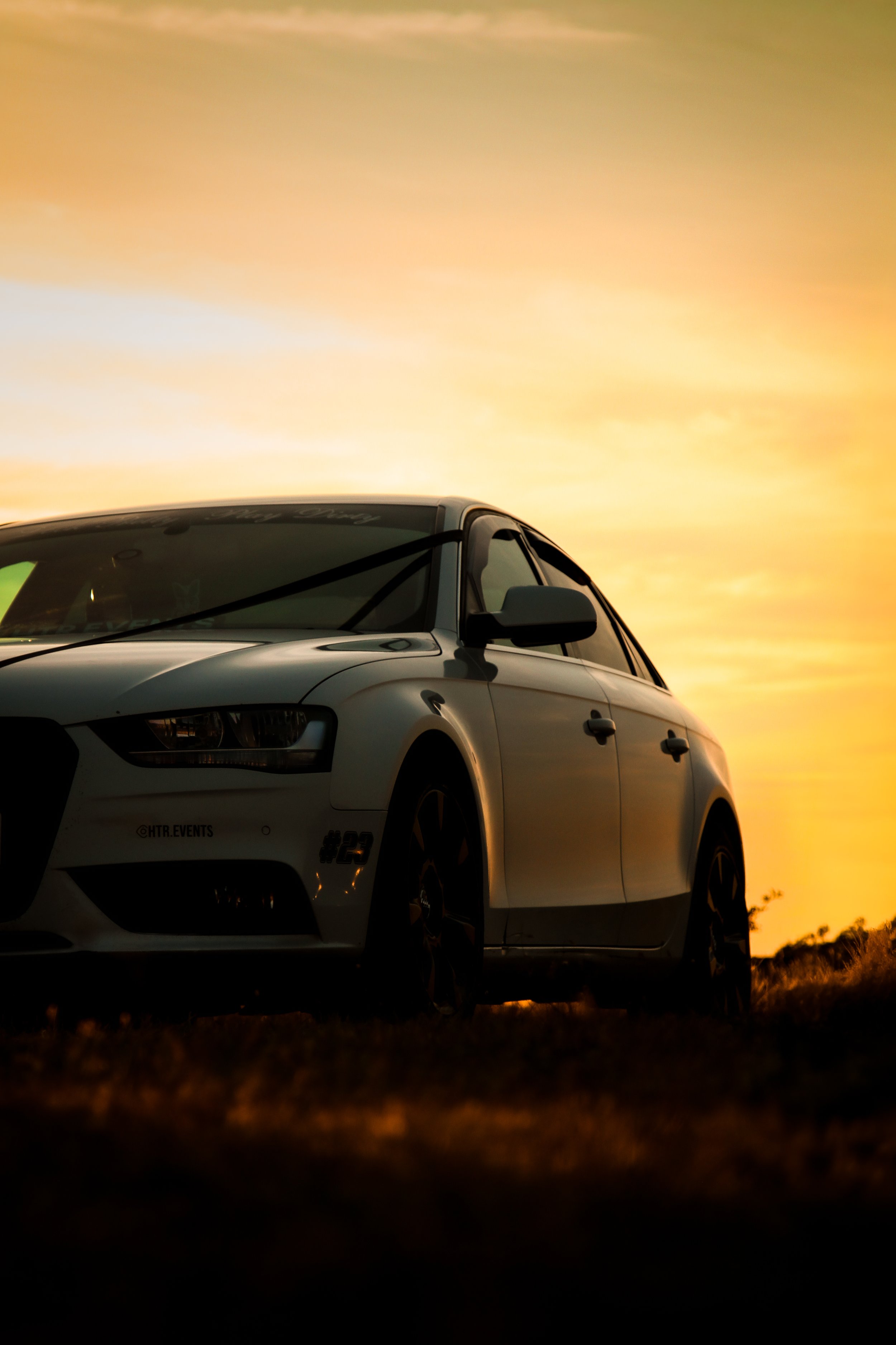A white car parked outdoors during sunset with an orange and yellow sky in the background.