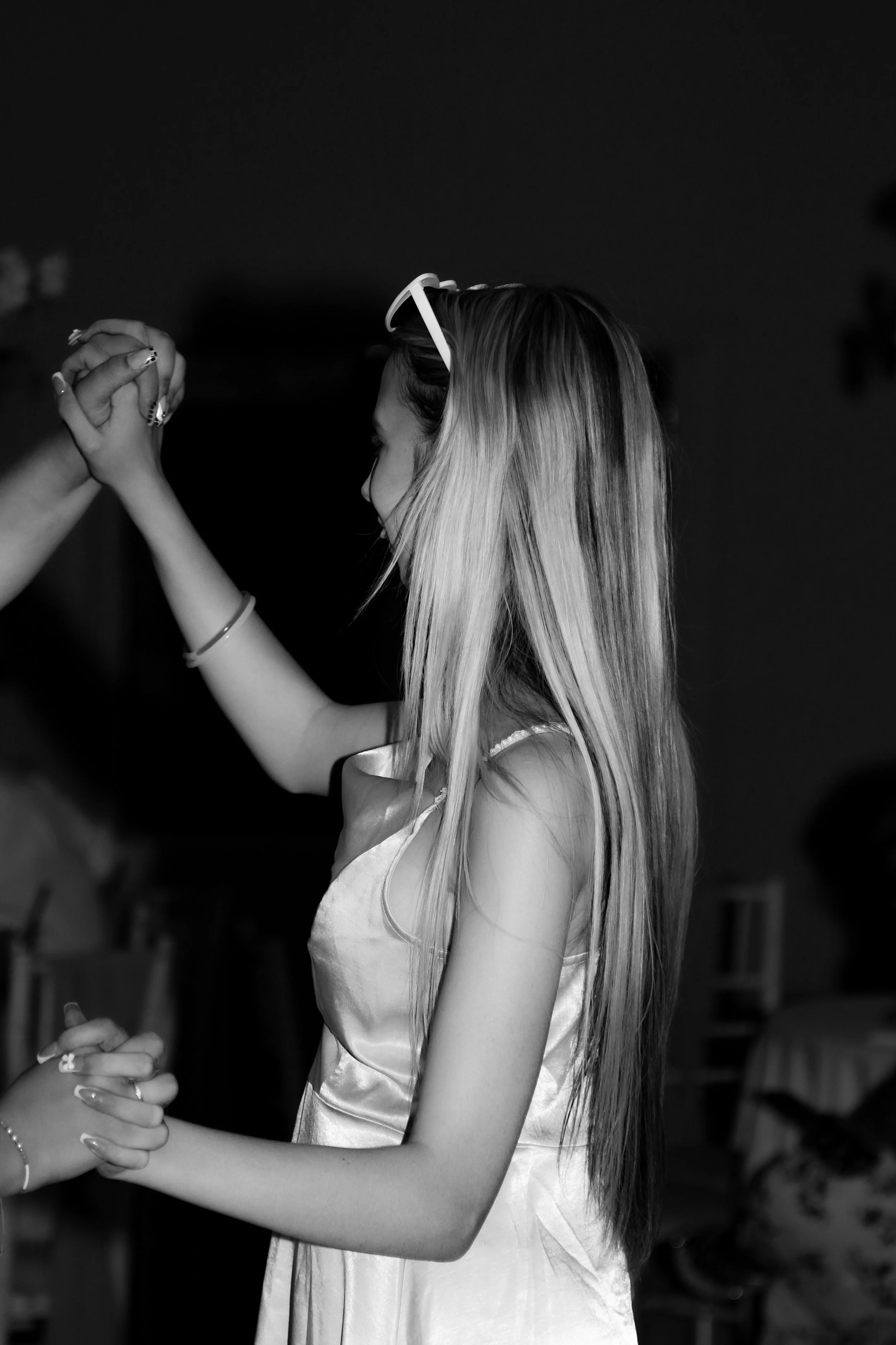 A young woman with long, blonde hair wearing a satin dress and glasses on her head is holding hands with another person in an indoor setting.