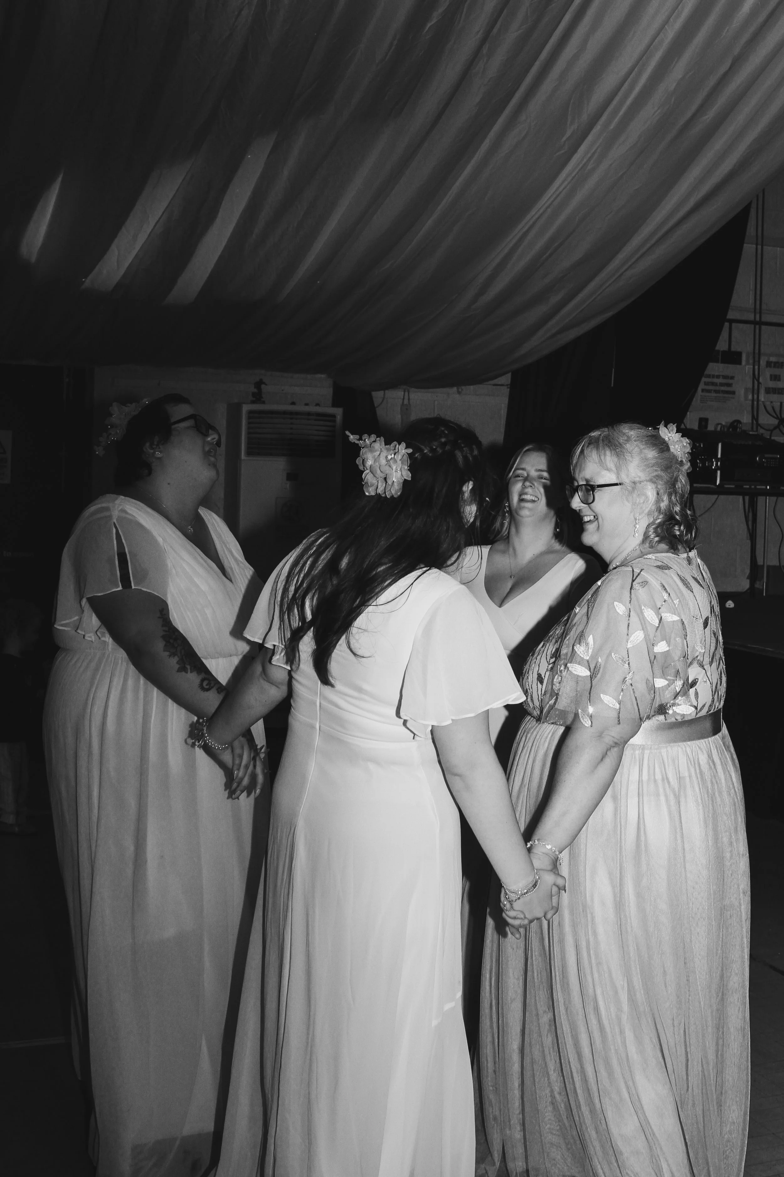 Group of women in light-colored dresses holding hands, appearing to exchange wedding vows in a dimly lit indoor setting with draped ceiling.
