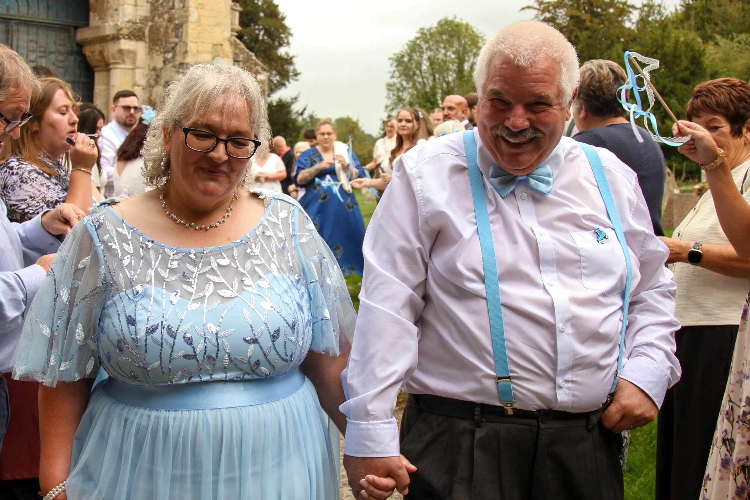 A group of people dressed in formal attire, holding hands, outdoors near an old stone building with trees in the background, celebrating a wedding or special event.