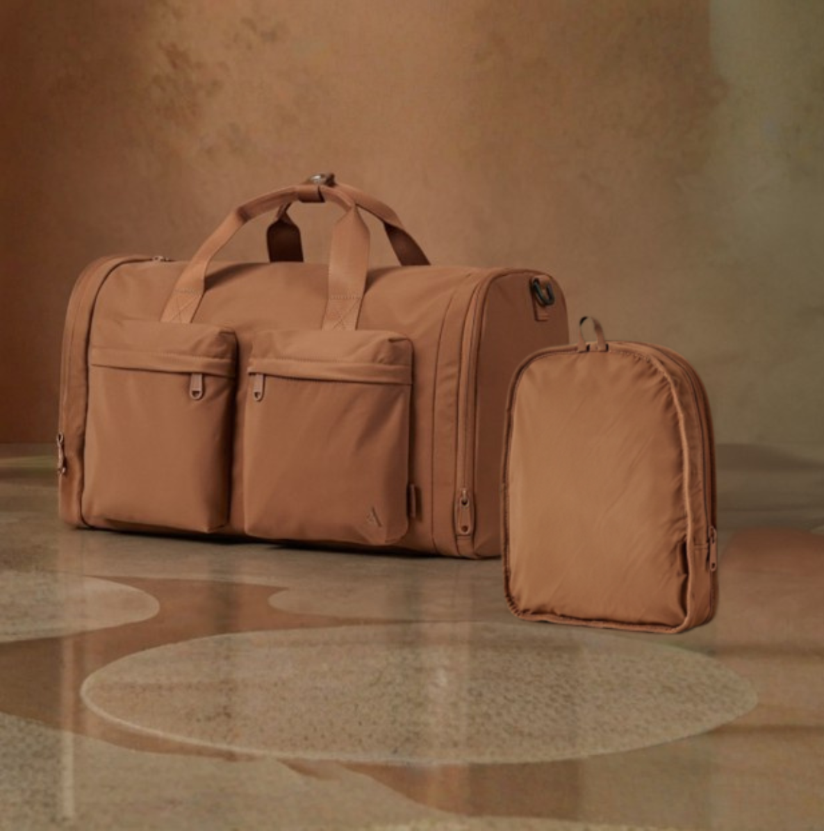 Beige travel duffel bag with two visible front zip compartments, folded into small carry bag on a wooden floor with a brown wall background.