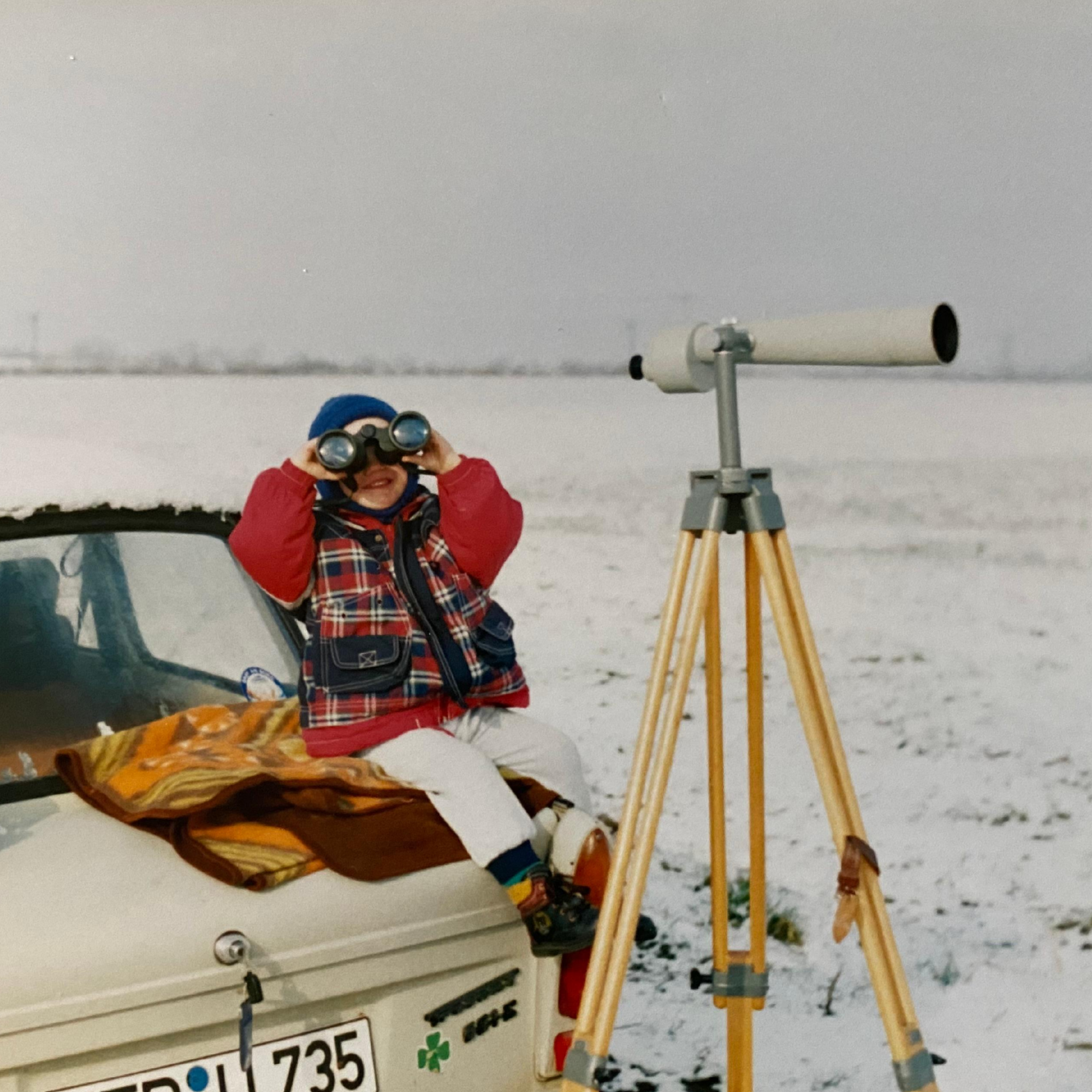 A person dressed in winter clothing is looking through binoculars at a snowy landscape. There is a large telescope on a tripod nearby and some vehicle parts below the image.