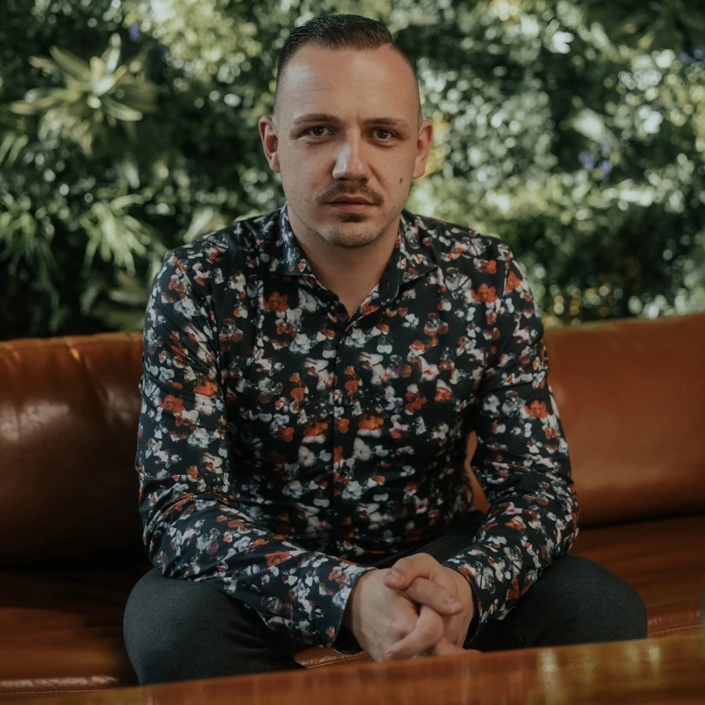 A man with slicked-back hair and a mustache, wearing a black floral shirt, sitting with arms crossed on a wooden table outdoors surrounded by green foliage.