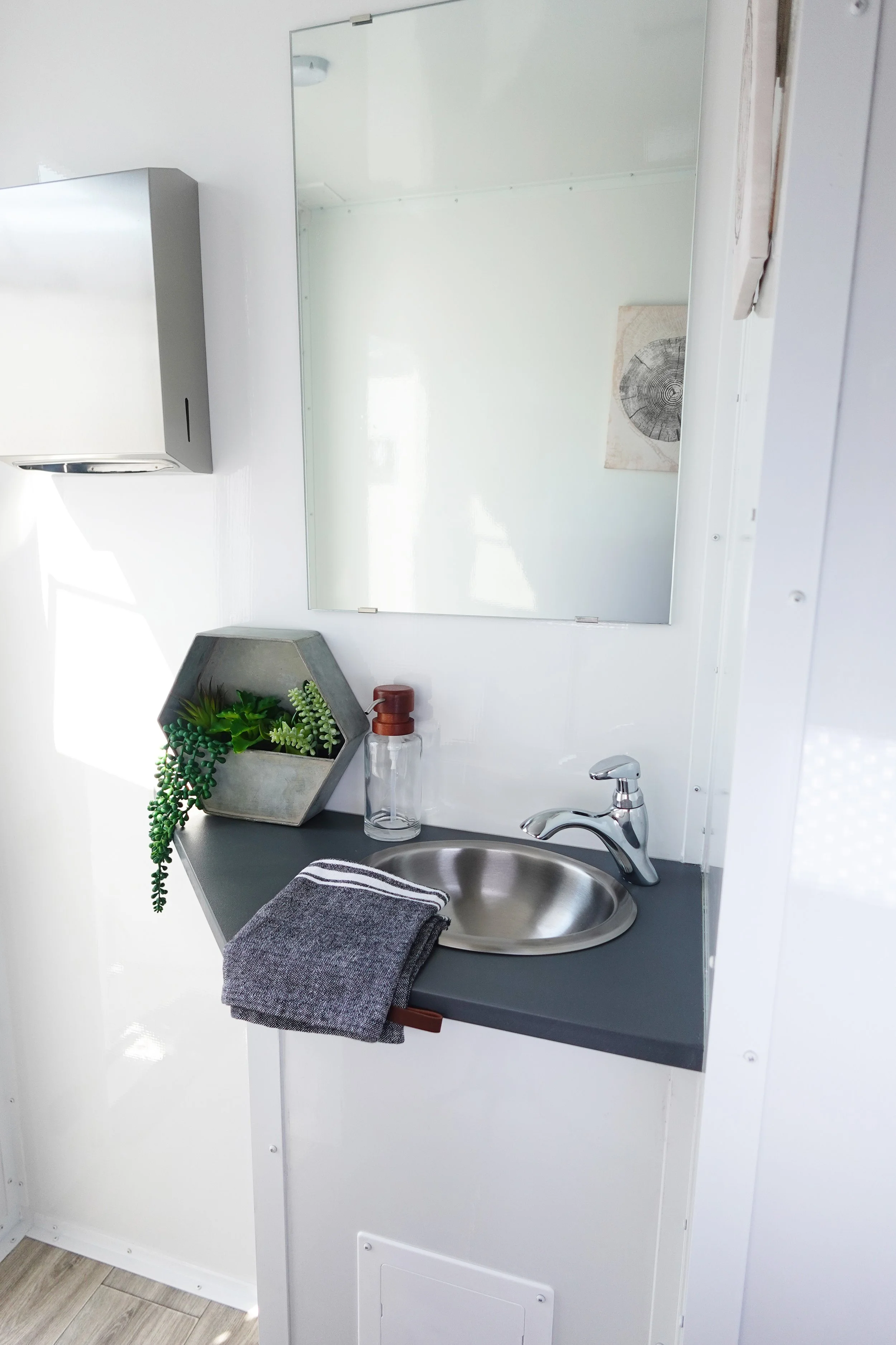 Small bathroom sink area with a round stainless steel sink, faucet, and a gray countertop. A folded dark gray towel is draped over the edge of the countertop. Above, a large rectangular mirror reflects a white wall and part of a small piece of artwork or a design featuring a tree ring pattern. To the left of the mirror, there is a wall-mounted gray dispenser. On the countertop, there is a decorative hexagonal planter with green succulents and a clear soap dispenser with a brown pump.