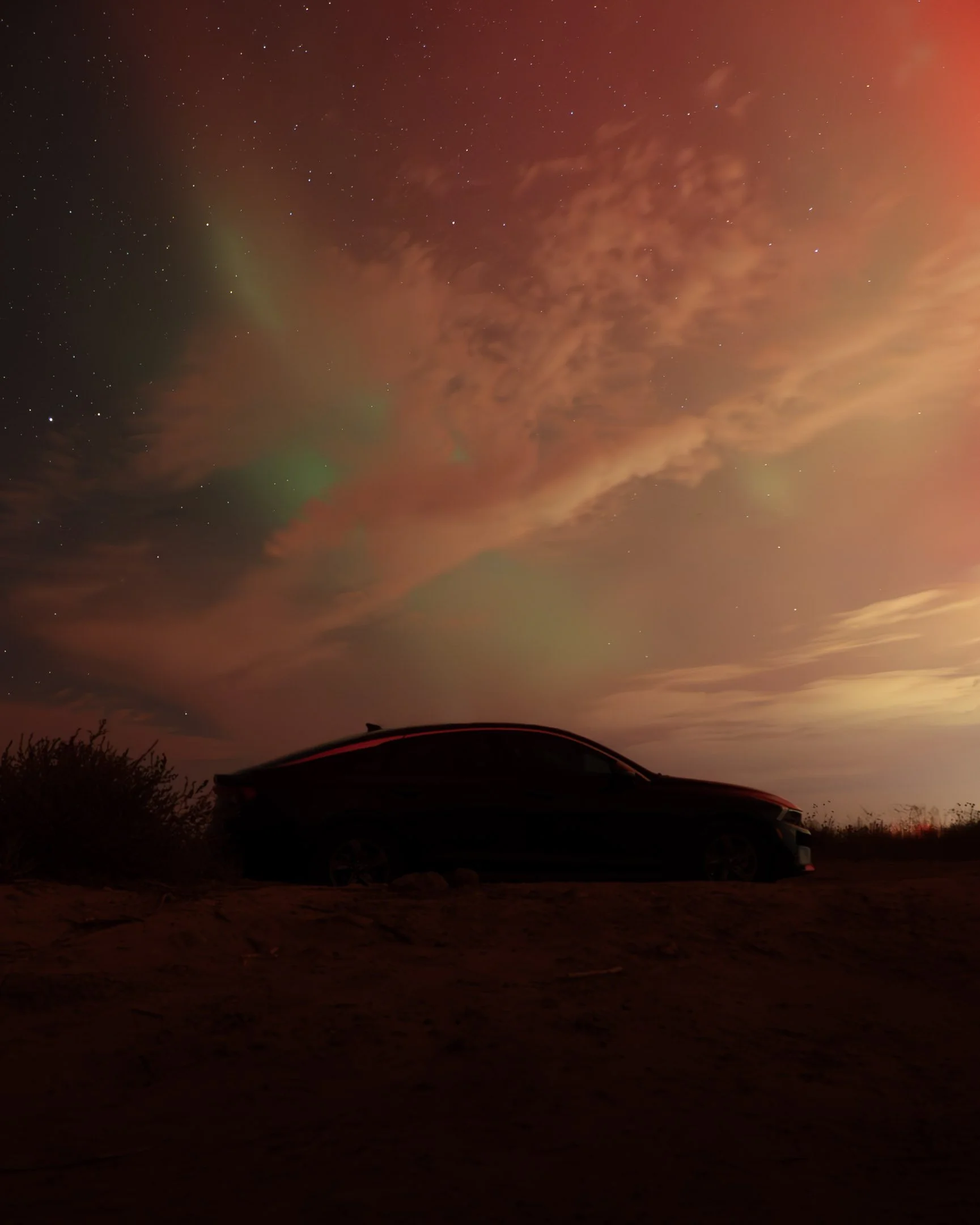 A car parked on a dirt patch under a night sky filled with stars and clouds, with trees silhouetted in the distance.