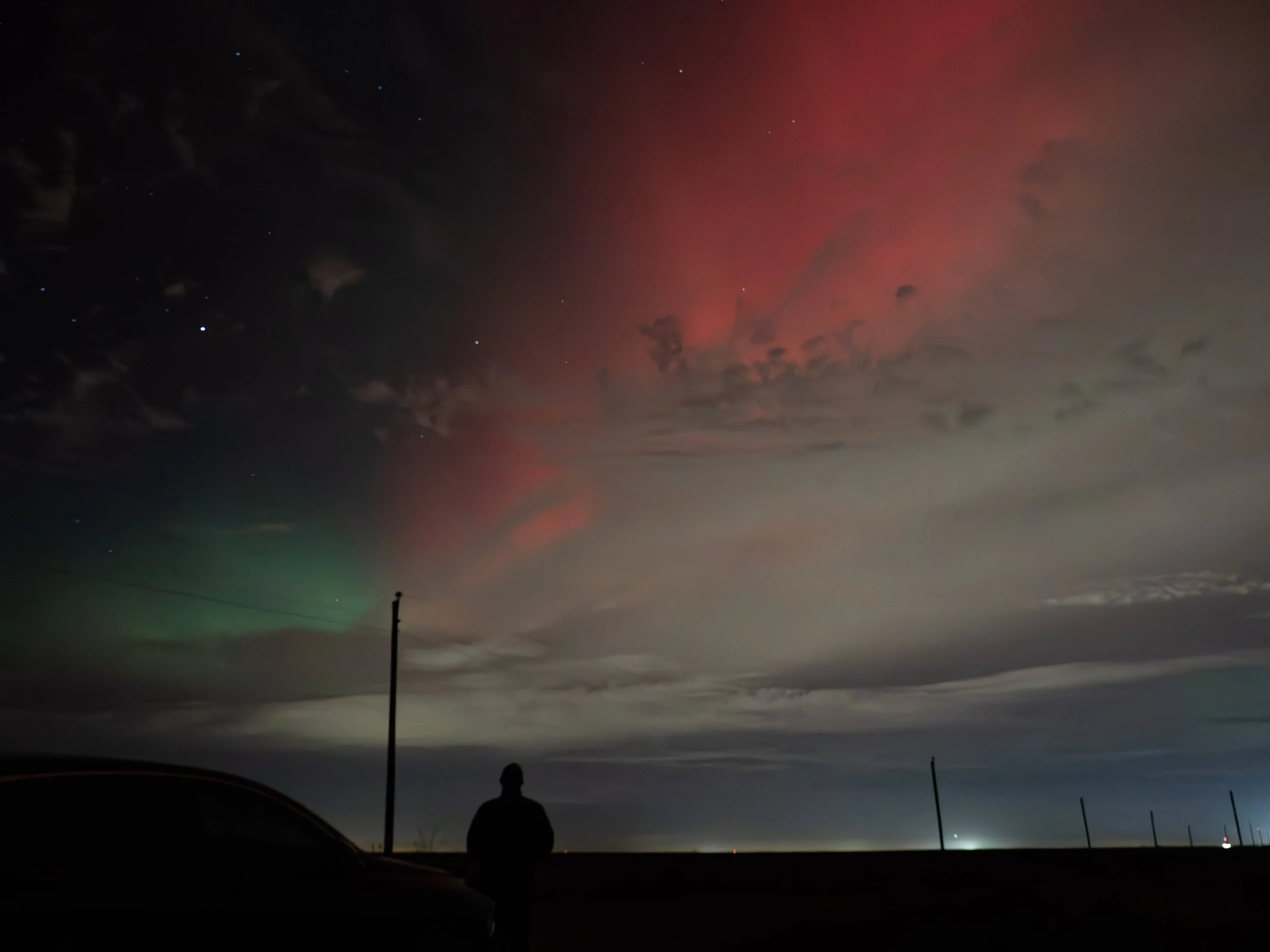 A person standing outdoors at night, silhouetted against the sky illuminated by a colorful display of the Northern Lights, with a few clouds and stars visible.