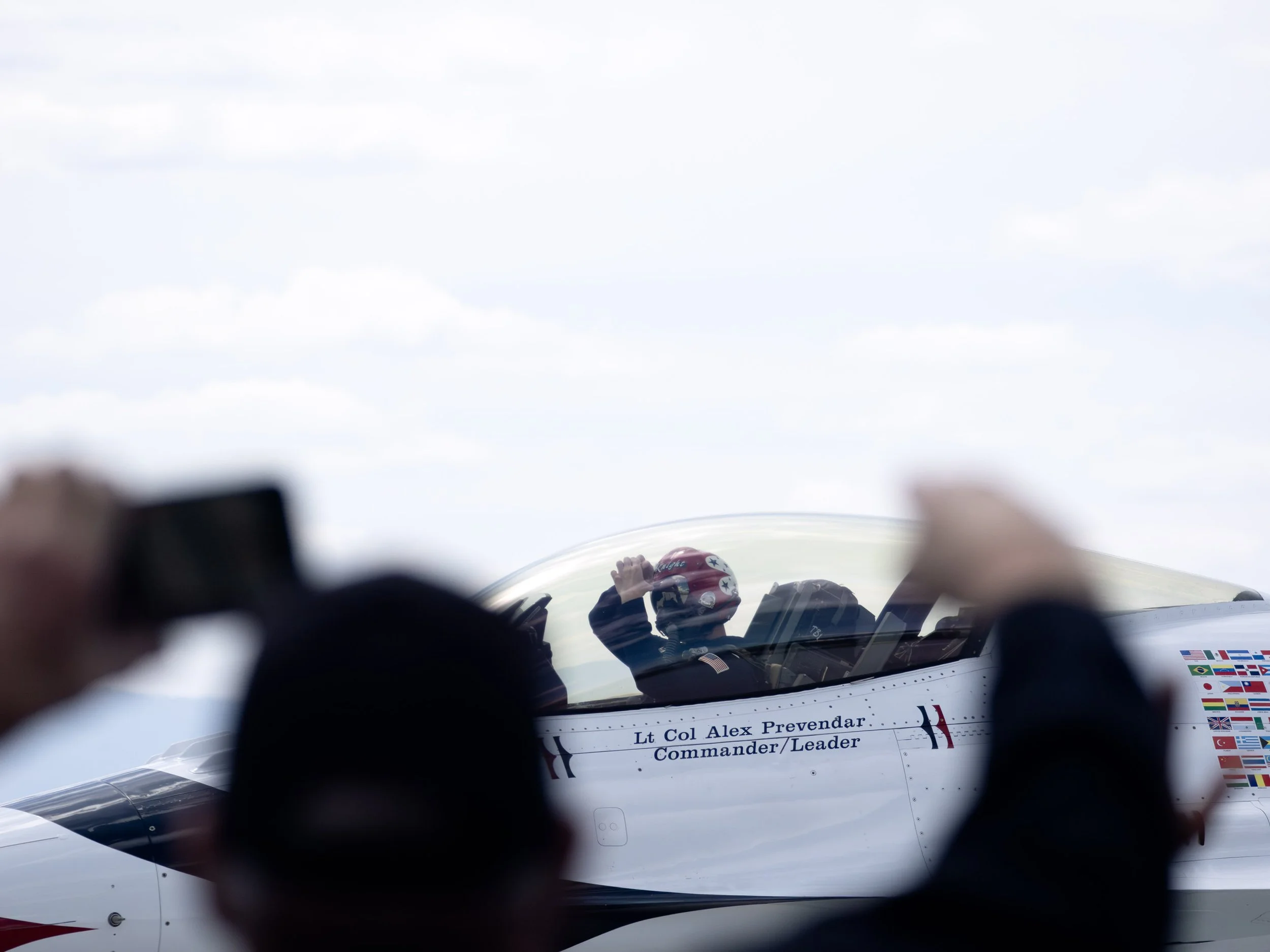 Close-up of a fighter jet cockpit, with a pilot wearing a helmet, reflections on the cockpit canopy, and a sign reading 'Lt Col Alex Prevendar Commander/Leader' visible on the side of the jet.