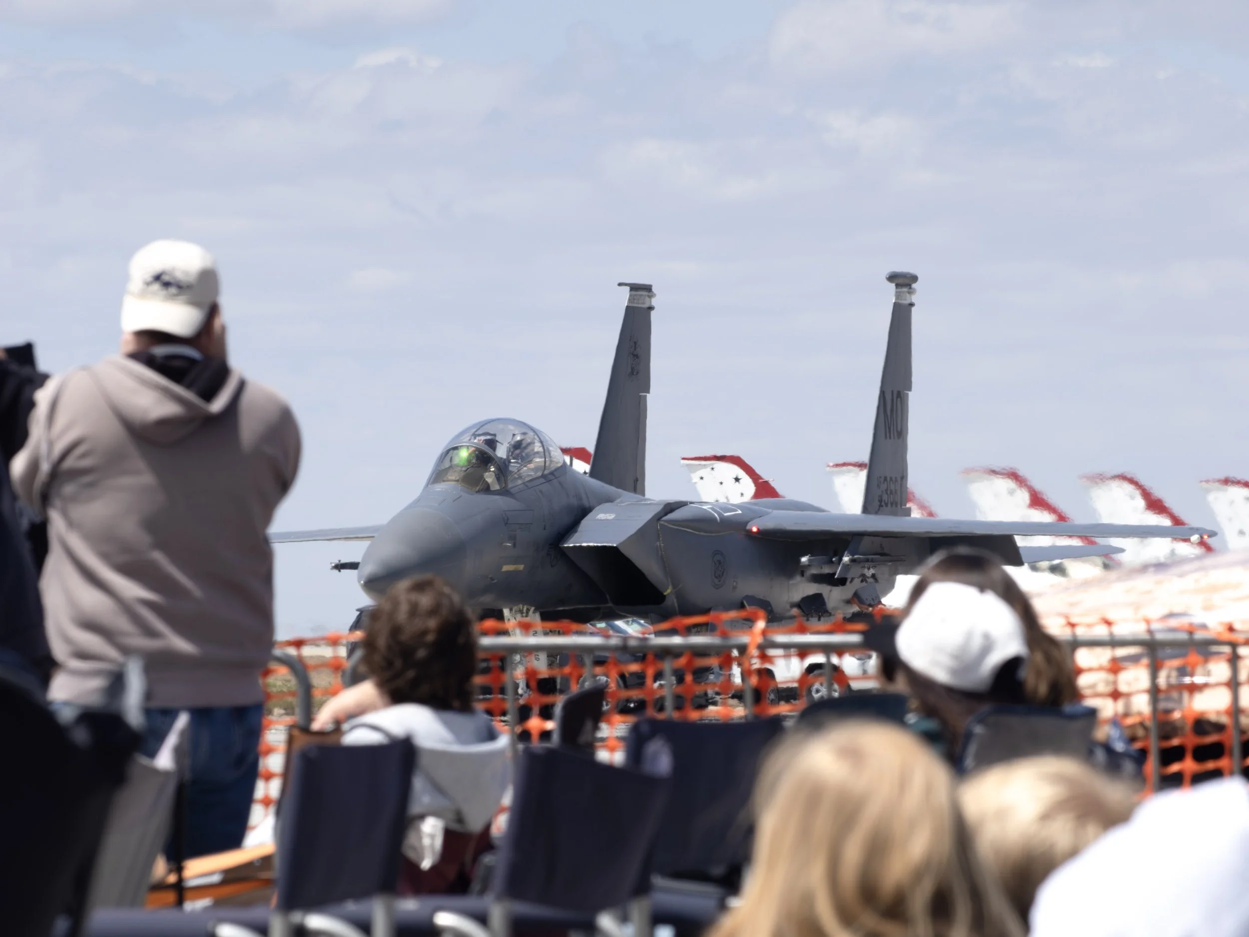 A crowd of people watching a military aircraft on an airshow. The aircraft is an F-15 fighter jet with afterburners producing red exhaust and a pilot visible in the cockpit.