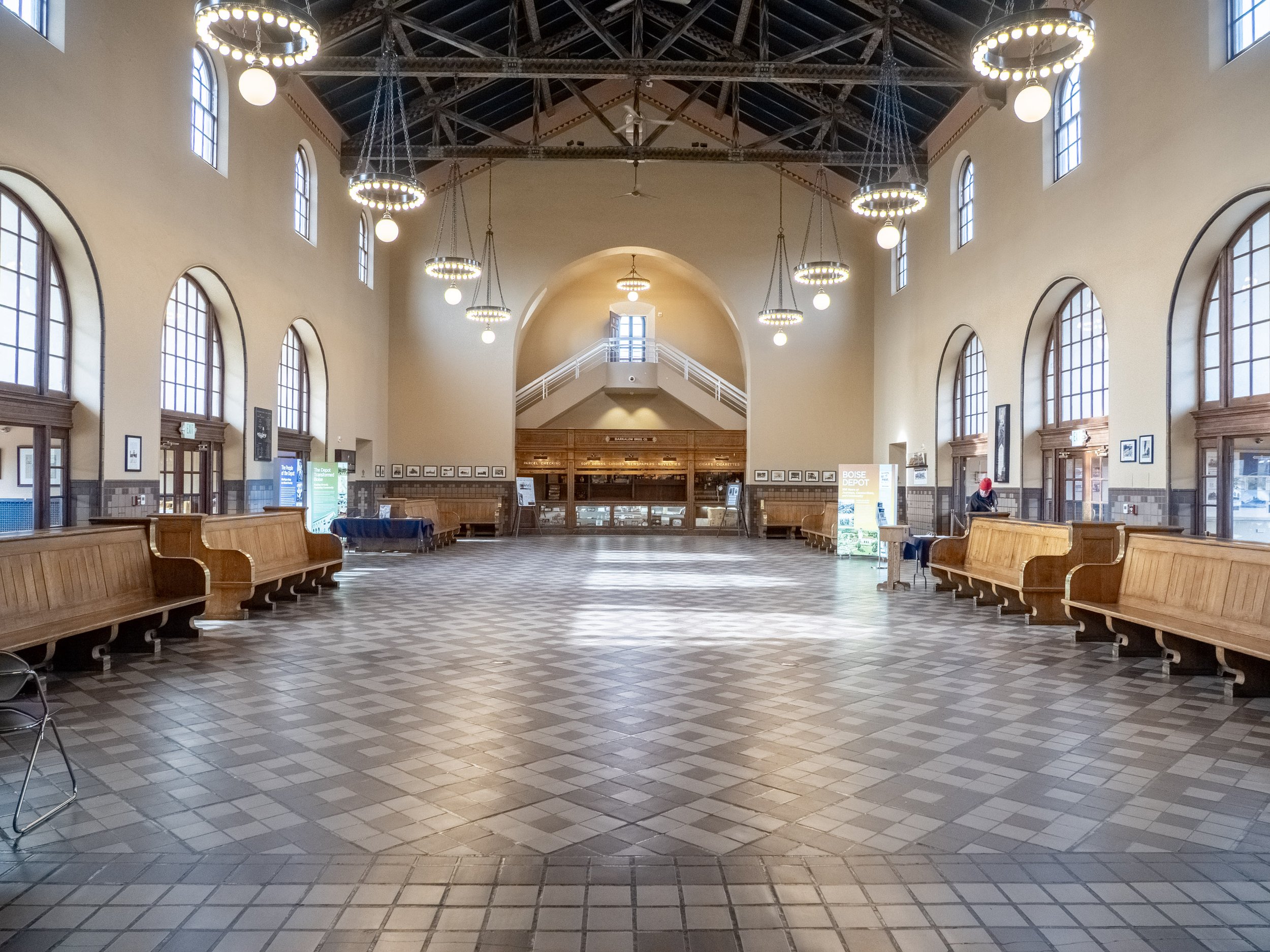 Interior of a spacious train station or waiting hall with high arched windows, wooden benches, and vintage chandeliers hanging from the ceiling.