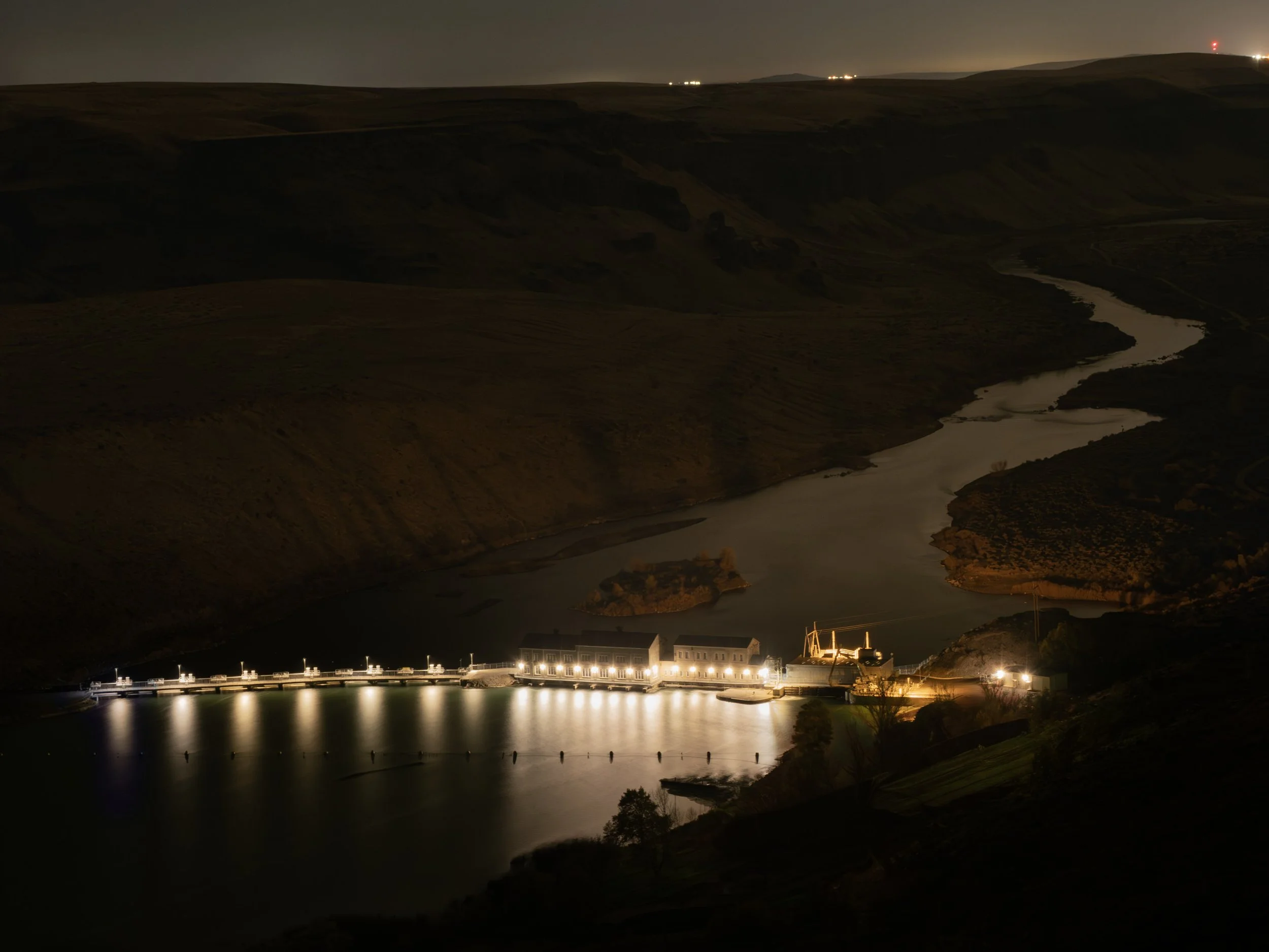 Night view of a river with reflections of Swan Falls Dam, hills in the background, and buildings with lights along the shoreline.