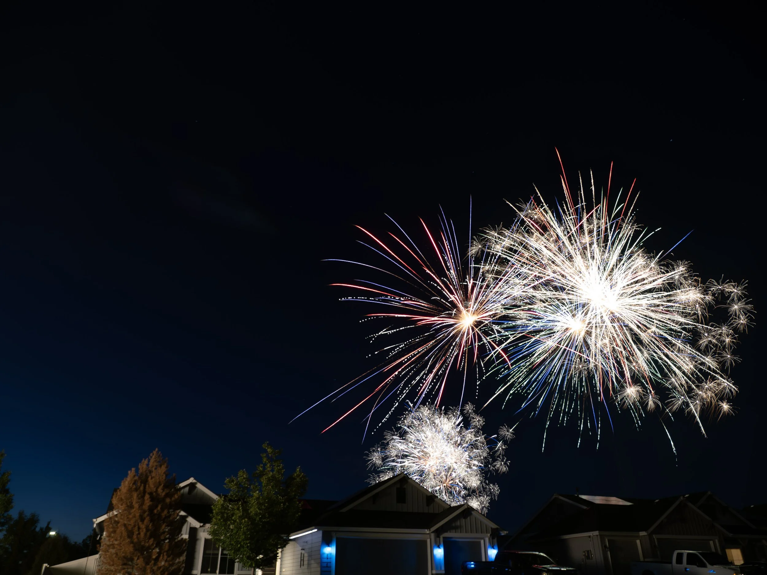 Colorful fireworks display lighting up the night sky above houses and trees.
