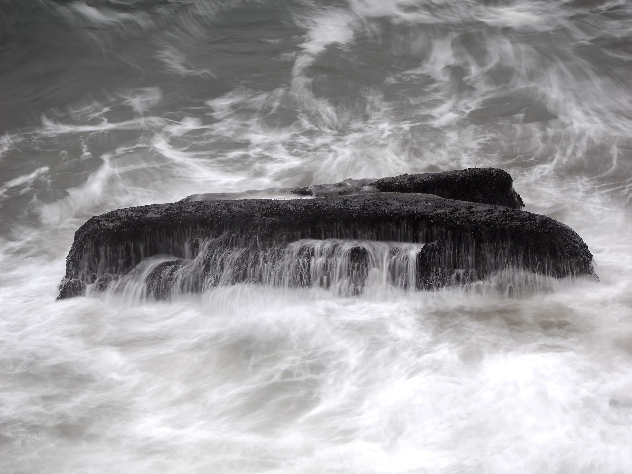 Waves crashing against dark rocks at the shoreline with white foam and swirling water.