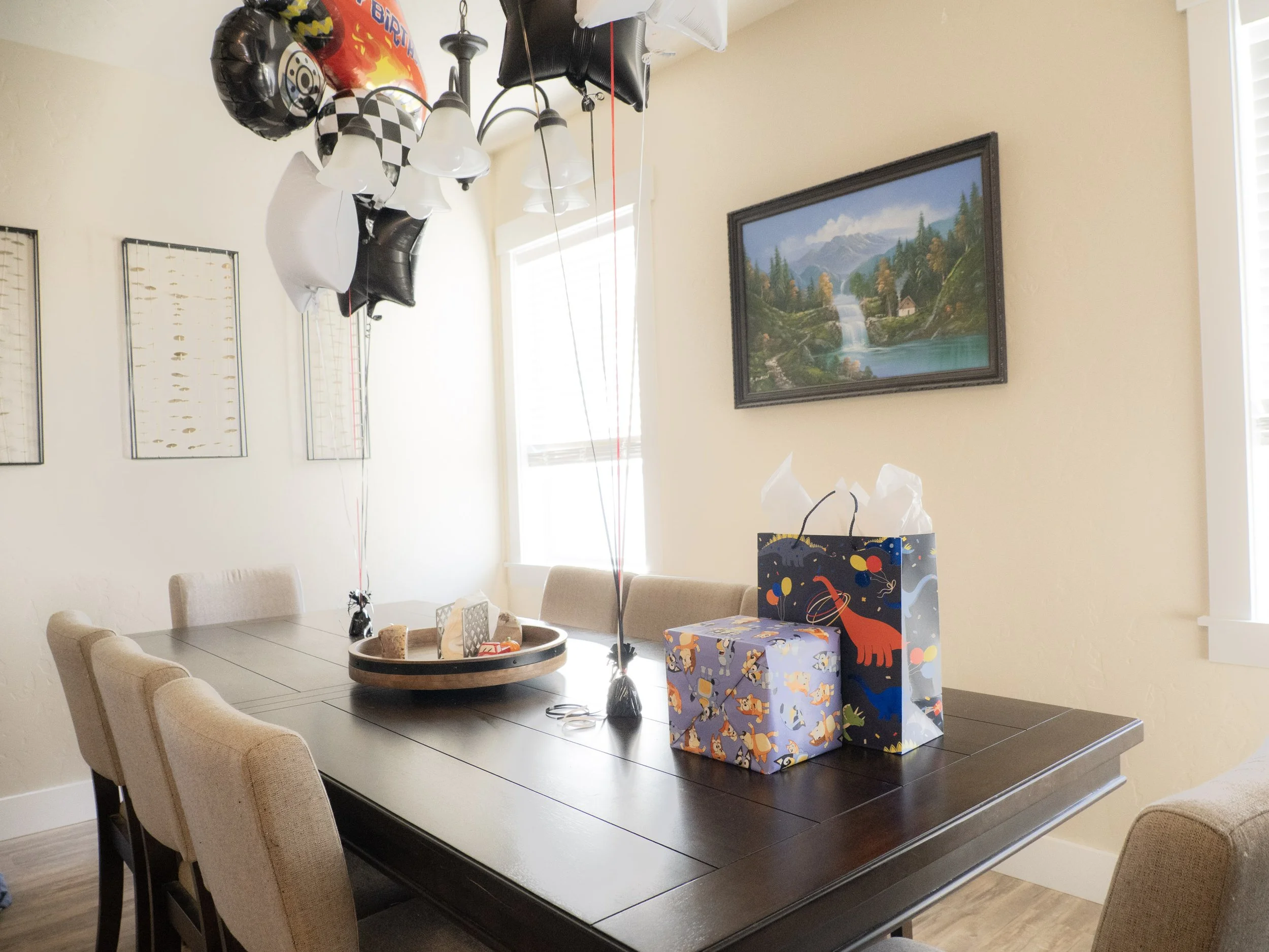 A dining room with a dark wood table and beige chairs, decorated for a celebration with balloons, gift bags, and a framed landscape painting on the wall.
