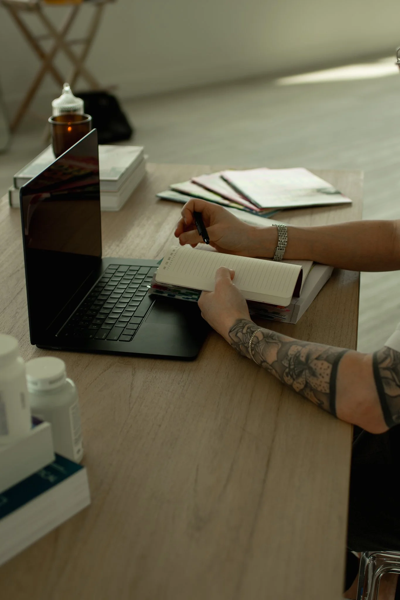 A person with tattoos on their arms writing in a notepad at a wooden desk. The desk has a laptop, a stack of magazines or notebooks, and a few bottles. The background shows a blurred room with some furniture and a floor made of light-colored wood.
