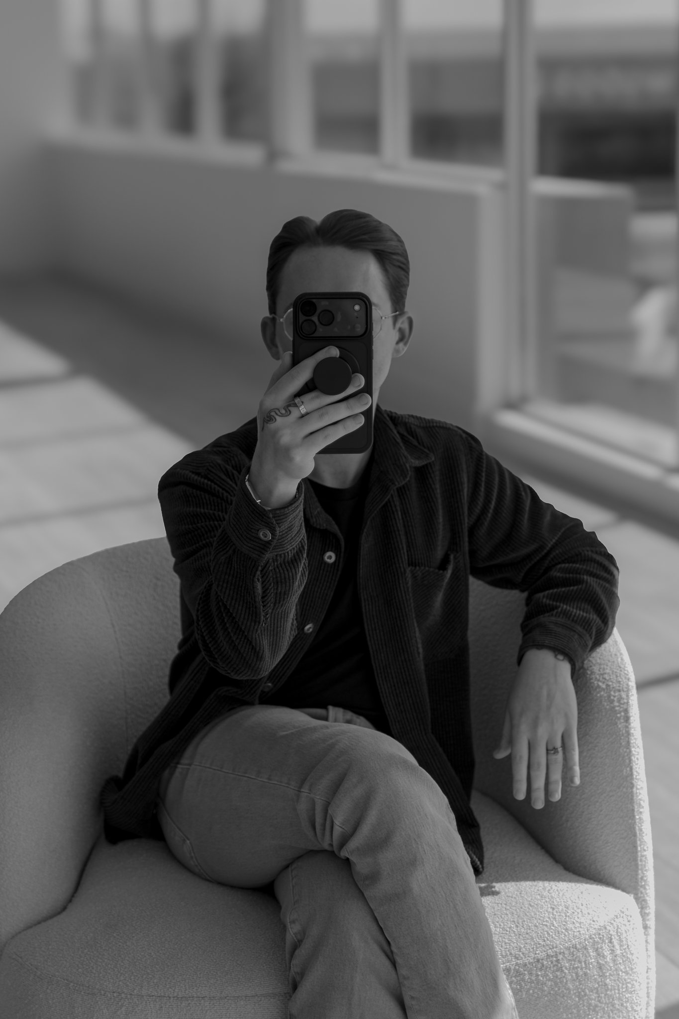 Black and white photo of a person sitting on a curved couch, taking a mirror selfie with a smartphone. The person's face is hidden behind the phone, wearing glasses, a ring, and a ring on the other hand's finger. The individual has styled hair and is dressed in casual clothing, including a jacket and pants. The background features a blurred modern interior with large windows and furniture.