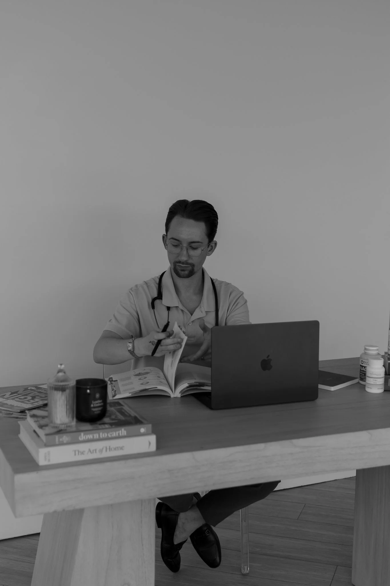 A man with glasses, a mustache, and a beard, wearing a medical coat with a stethoscope, is sitting at a desk and reading a book. The desk has a laptop, medicine bottles, and books on it.