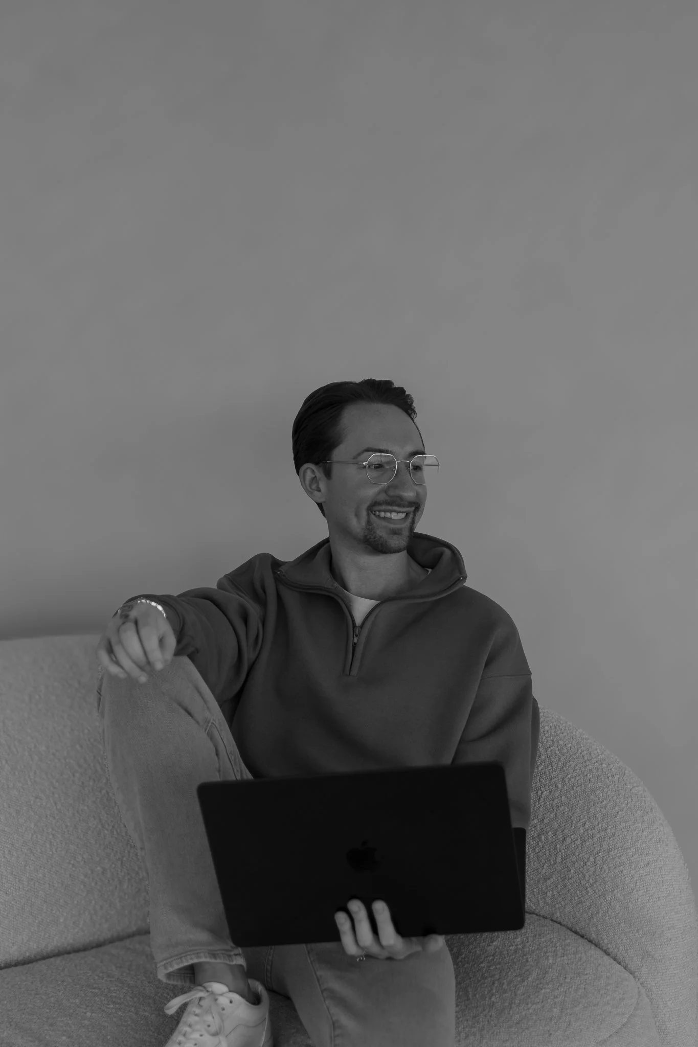 A man sitting on a couch, smiling, holding a laptop in his lap, wearing glasses, a zip-up hoodie, and sneakers.