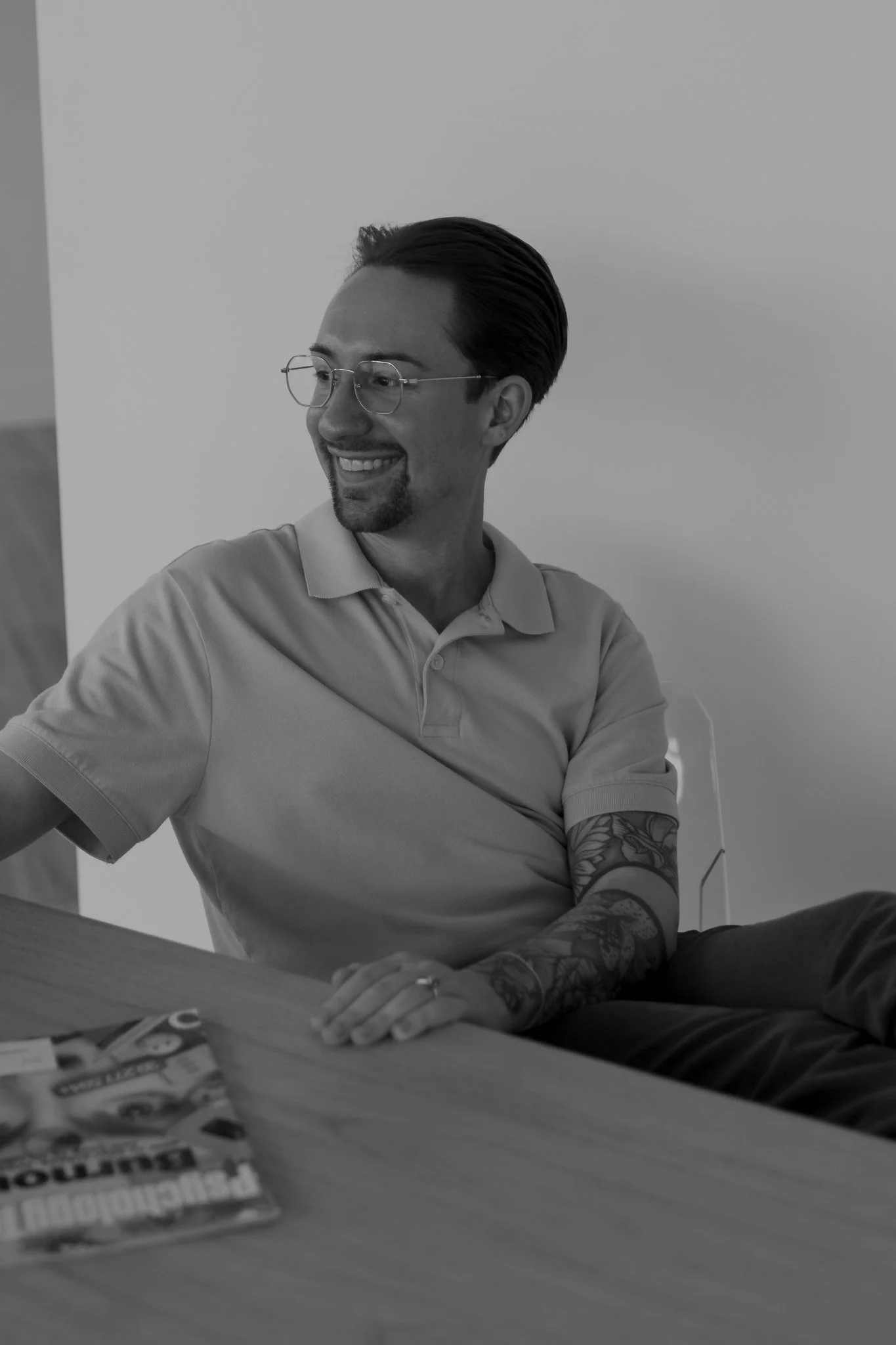 A man with glasses and a beard, wearing a light-colored polo shirt, smiling and sitting at a table. There are magazines or newspapers on the table in front of him, and his left arm is resting on the table, revealing tattoos on his forearm.