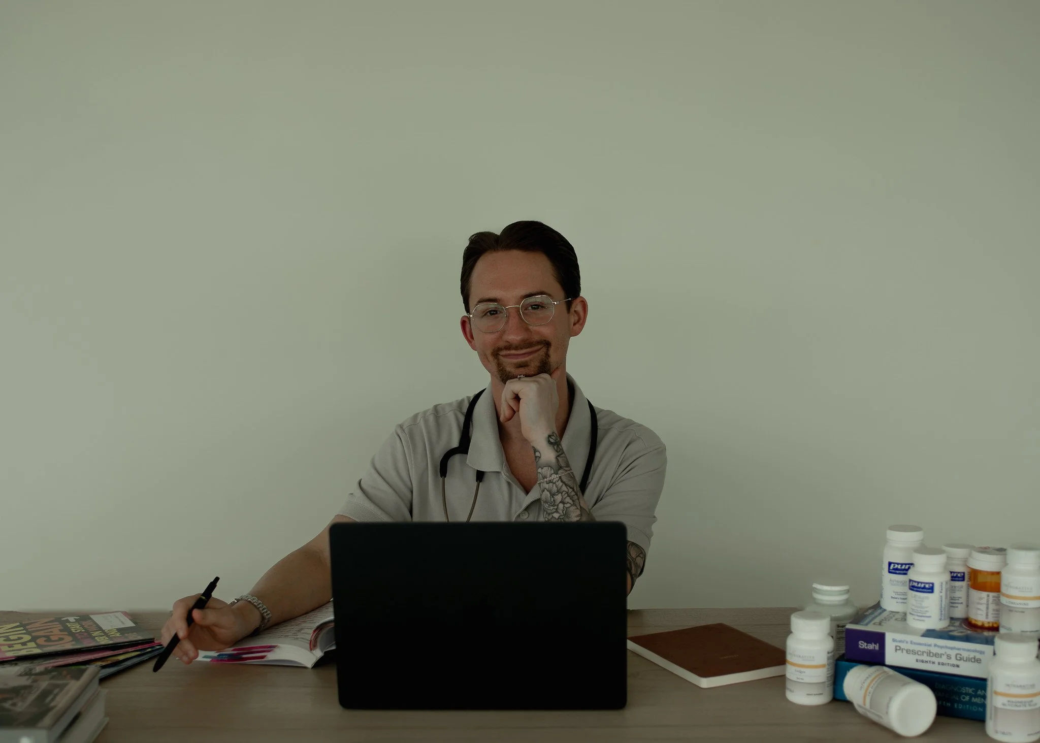 A male doctor with glasses and tattoos, sitting at a desk in a medical office, smiling and resting his chin on his hand, surrounded by medication bottles, a laptop, and magazines.